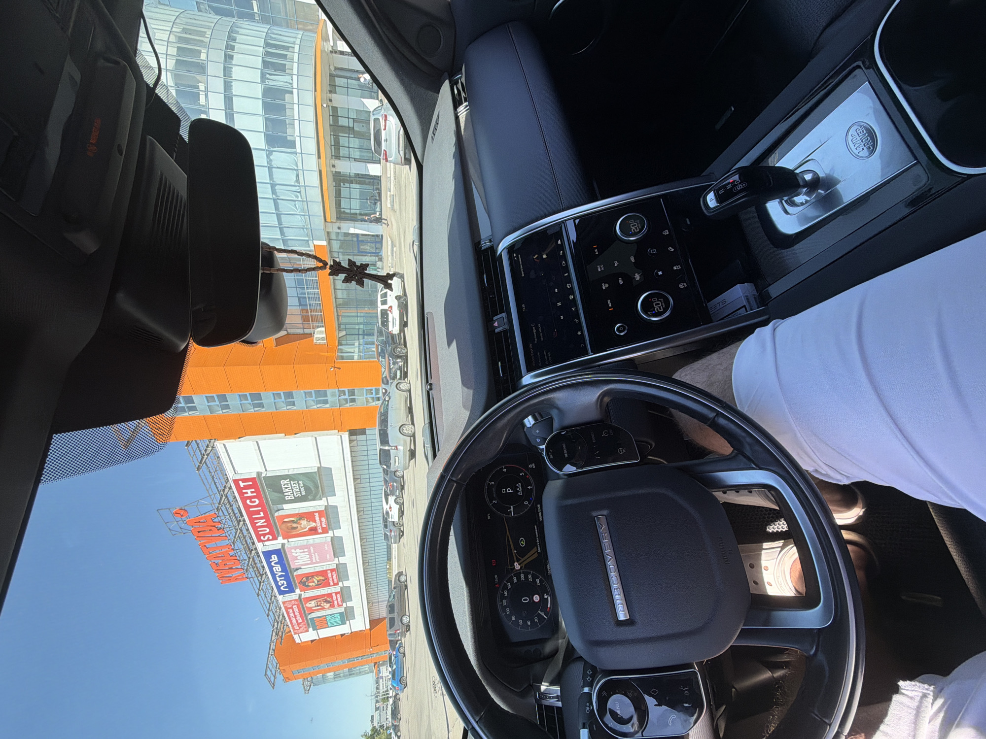The image provides a first-person perspective from the driver's seat of a Land Rover Discovery vehicle, looking out through the front windshield on a clear, sunny day in Krasnoyarsk, Russia.
**Vehicle Interior:**
The steering wheel, emblazoned with "DISCOVERY," is prominent in the foreground. The digital instrument cluster behind it displays a speed of 0, an external temperature of 30°C, the time as 13:20, and the date as 08/06/2026. Text on the display reads "Дорога без названия" (Road without a name). Below the steering wheel, a person's legs are visible, clad in white pants and light-colored sneakers. The center console features a touchscreen display with climate controls set to 20.0°C, and below it, a gear selector in "P" (Park) mode, with the "LAND ROVER" logo embossed on its surrounding trim. A small stack of white papers or cards is tucked into a slot near the gear selector. A rearview mirror hangs from the ceiling, from which a wooden crucifix pendant dangles. Above the mirror, a module containing a red "SOS" button is visible.
**Exterior Environment:**
Through the windshield, a large, multi-story building with a modern design is visible, primarily orange and white with numerous windows. This building is identified as the "KUBATURA" (КУБАТУРА) shopping and business center, with its name displayed in large orange letters on a roof structure. Below the main sign, other illuminated retail signs are visible, including "Л'ЭТУАЛЬ" (L'Etoile), "SUNLIGHT," "Hoff," and "BAKER STREET." To the right of the main building, another modern structure with a curved glass facade is partially visible. A paved parking lot stretches in front of the buildings, occupied by multiple vehicles, including a silver SUV, a dark SUV, a white SUV, and a blue hatchback. Several individuals are seen walking in the distance near the entrances of the buildings. The sky is clear and blue, indicating bright daylight.