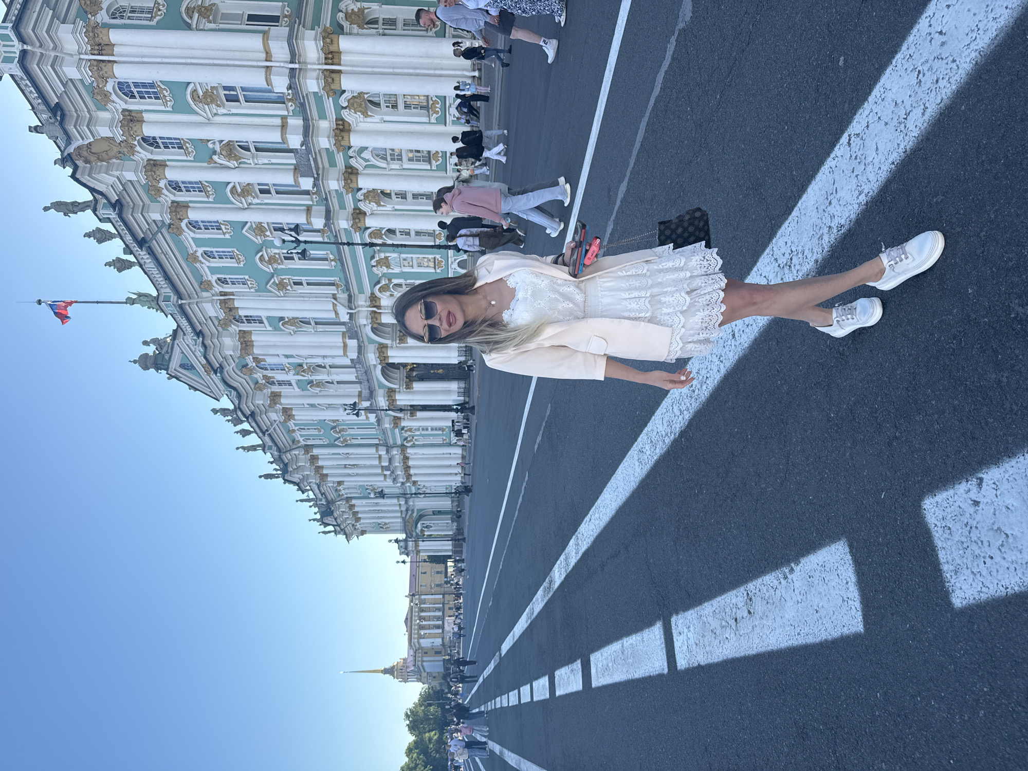 In Saint Petersburg, Russia, a woman stands on Palace Square, her gaze directed towards the left. She is wearing a cream-colored blazer over a white lace dress and white sneakers. She carries a dark patterned bag in her right hand and two phones in her left. Behind her, the grand facade of the Winter Palace, a former imperial residence and now part of the Hermitage Museum, stretches across the background. The building is characterized by its turquoise and white exterior, adorned with golden architectural details and numerous columns. Other people are visible walking on the square and in the distance. The sky is clear and blue, and the sun casts shadows on the asphalt of the vast square.