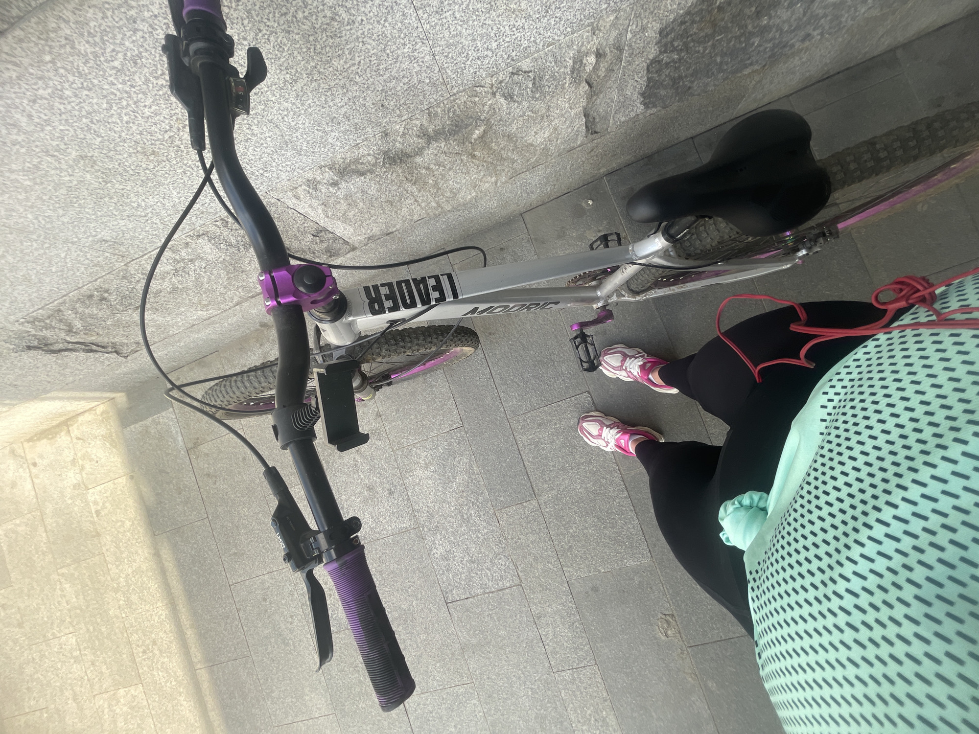 The image shows a Leader MOORC mountain bike, likely in Moscow, Russia, based on the urban paving stone visible. The bike is silver with purple accents on the handlebars, pedals, and possibly other components. A person wearing pink and white sneakers, black leggings, and a light blue, patterned shirt stands next to the bike. The angle suggests the photo was taken from an elevated position or by someone standing close to the subject. The background is composed of grey paving stones.