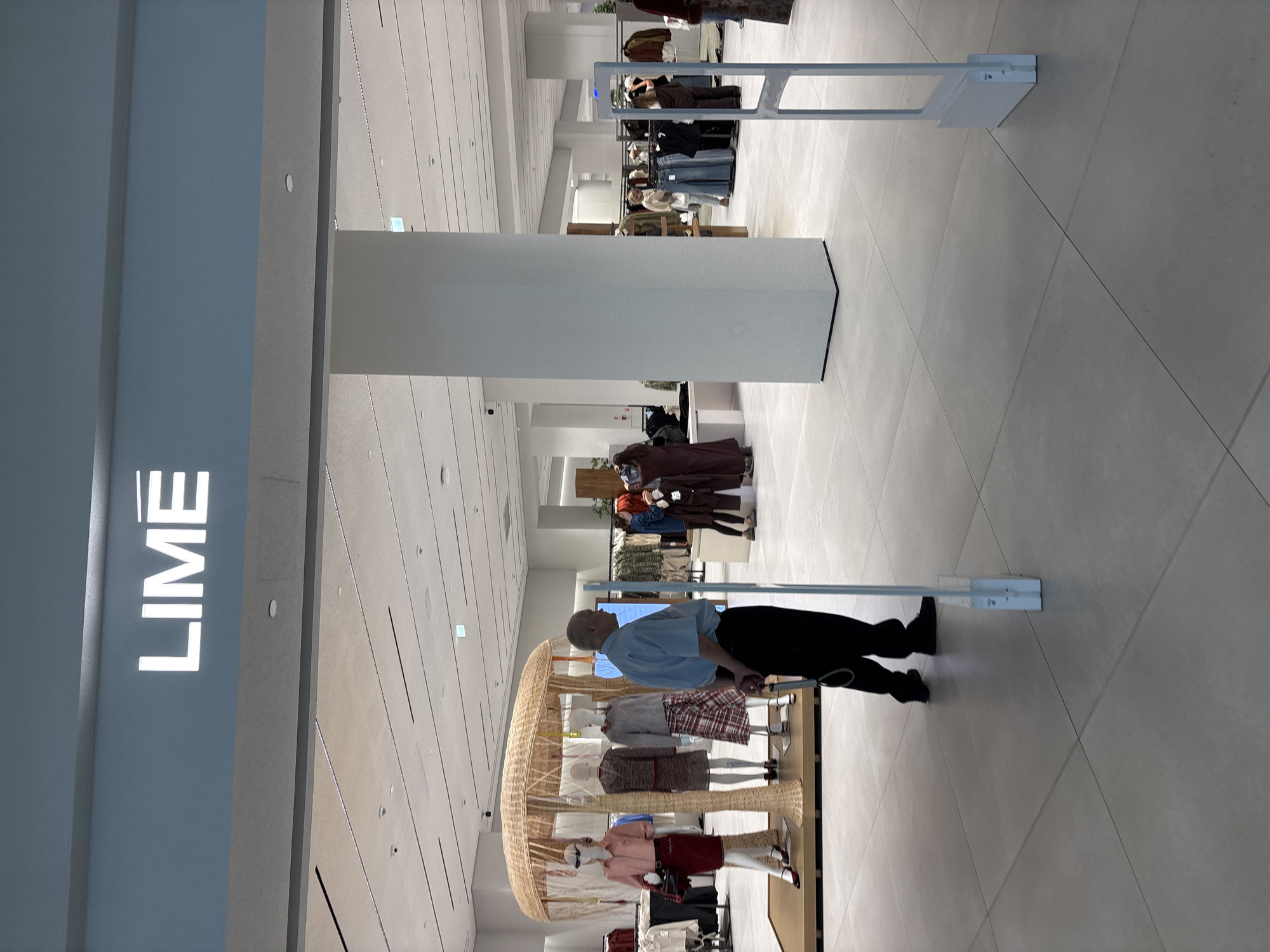 The image shows the interior of a brightly lit retail store, likely a clothing store. The prominent signage above the entrance reads "LIME" in large white letters against a light blue background. In the foreground, there are two white security gates, one on each side, that appear to be part of an entrance or exit system. A man wearing a light blue short-sleeved shirt and dark pants stands between the gates, facing away from the camera, with his hands clasped behind his back. He is partially obscured by a white pillar. To the left, a display features mannequins dressed in stylish clothing, including a pink shirt and a red skirt, and a tweed suit. Behind the display, racks of clothing are visible. Further into the store, other shoppers and displays of clothing can be seen. The flooring is light-colored and tiled. The overall environment suggests a modern and well-maintained commercial space. This scene is located within the city of Khimki, Russia.