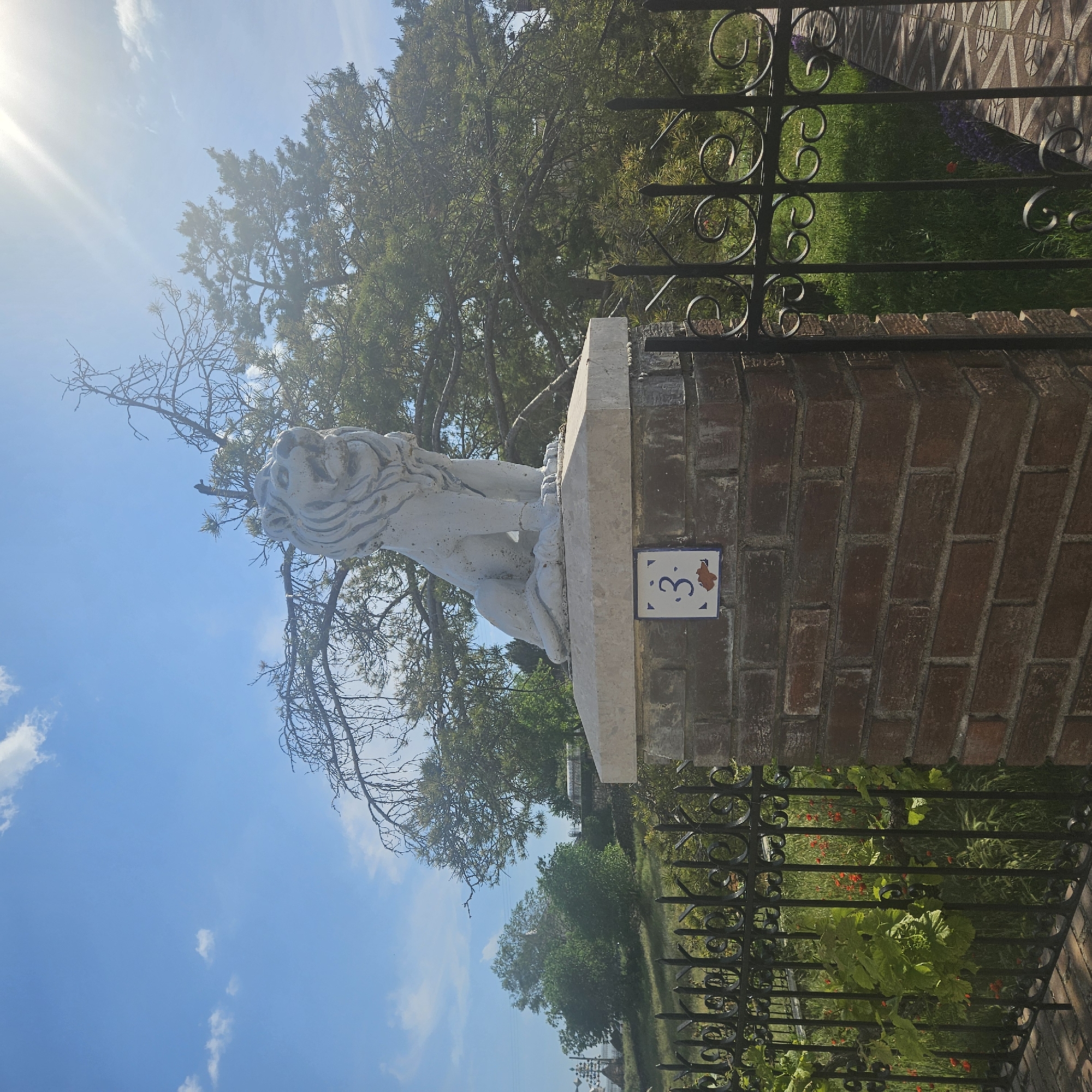 The image shows a stone lion statue perched on a brick pillar with a tile displaying the number "3". The pillar is adjacent to a black wrought-iron fence with decorative scrolls. Behind the fence, there is vegetation including grass, trees, and some plants with red flowers. The sky is blue with some clouds, and there is a bright glare from the sun. Based on the tile and the style of architecture, this image is from Algora, Spain.
