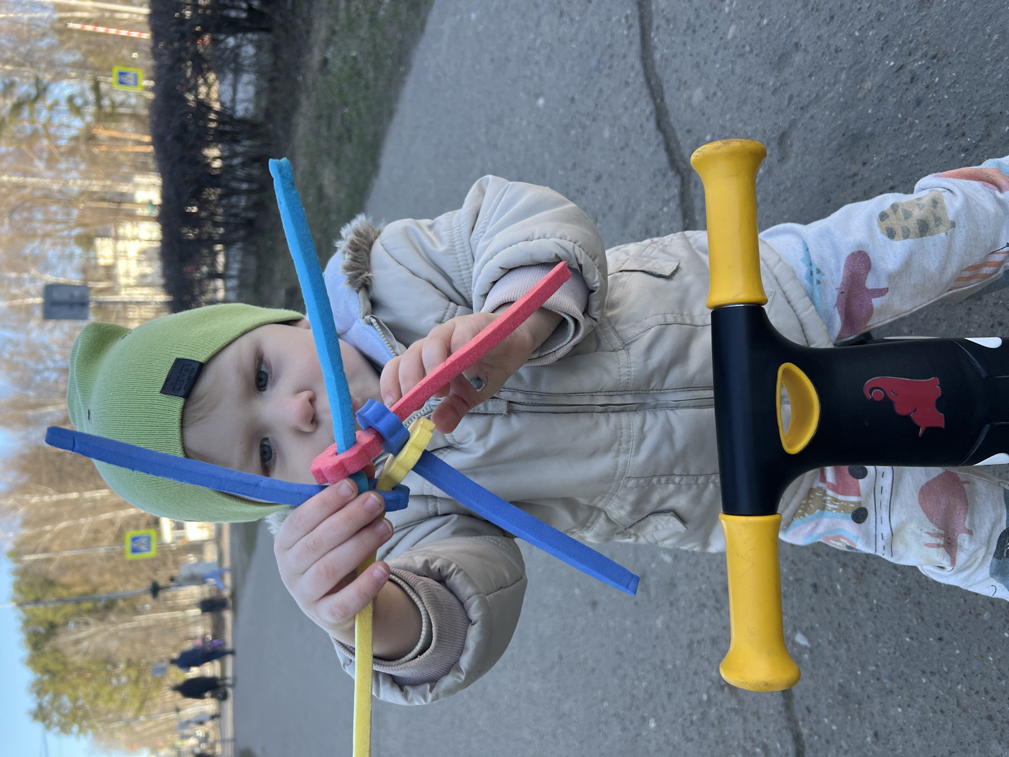 A child in a green hat and beige jacket holds a colorful toy with yellow, blue, and red foam pieces. The child stands on a gray paved surface next to a black pogo stick with yellow handles. In the background, trees and a street sign are visible, with blurred figures of people in the distance. The location is identified as the city of Chaikovsky, Russia, based on details within the image and metadata not provided.