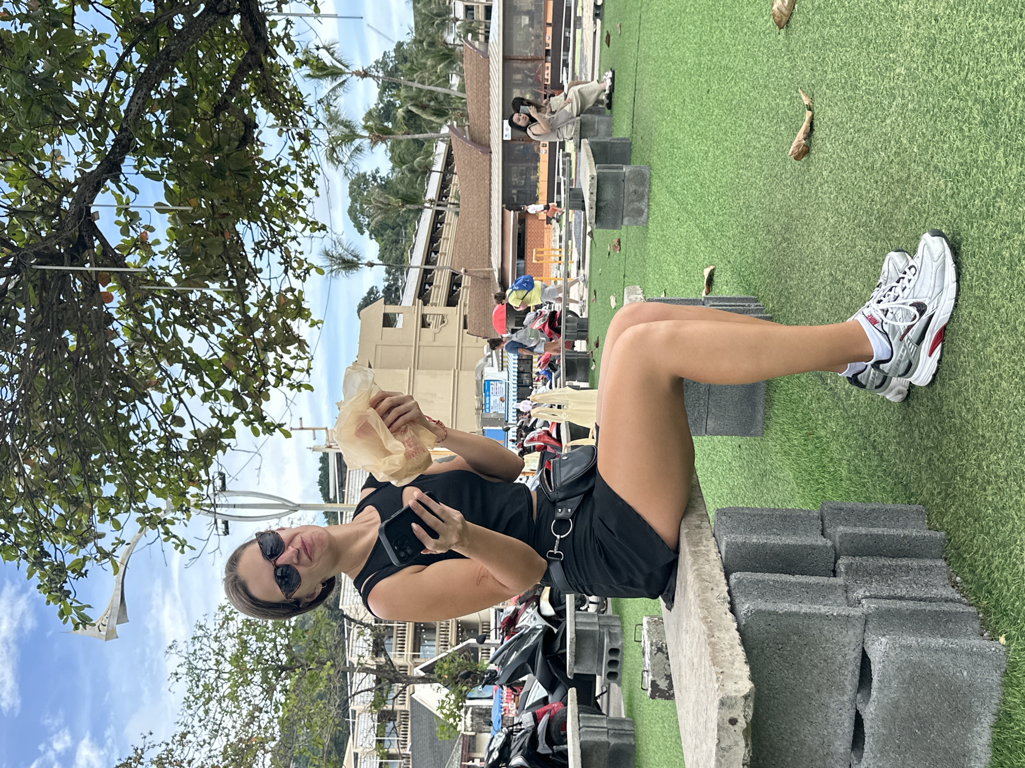 In Karon, Thailand, a young Caucasian woman wearing a black tank top, black mini-skirt, sunglasses, and white sneakers is seated on a concrete block bench. She holds a phone in her left hand and a wrapped food item in her right. She is positioned outdoors on a patch of artificial turf, with trees and buildings visible in the background. Other people are present in the background, including individuals walking, sitting on benches, and riding motorbikes. The scene is illuminated by natural daylight under a partially cloudy sky.