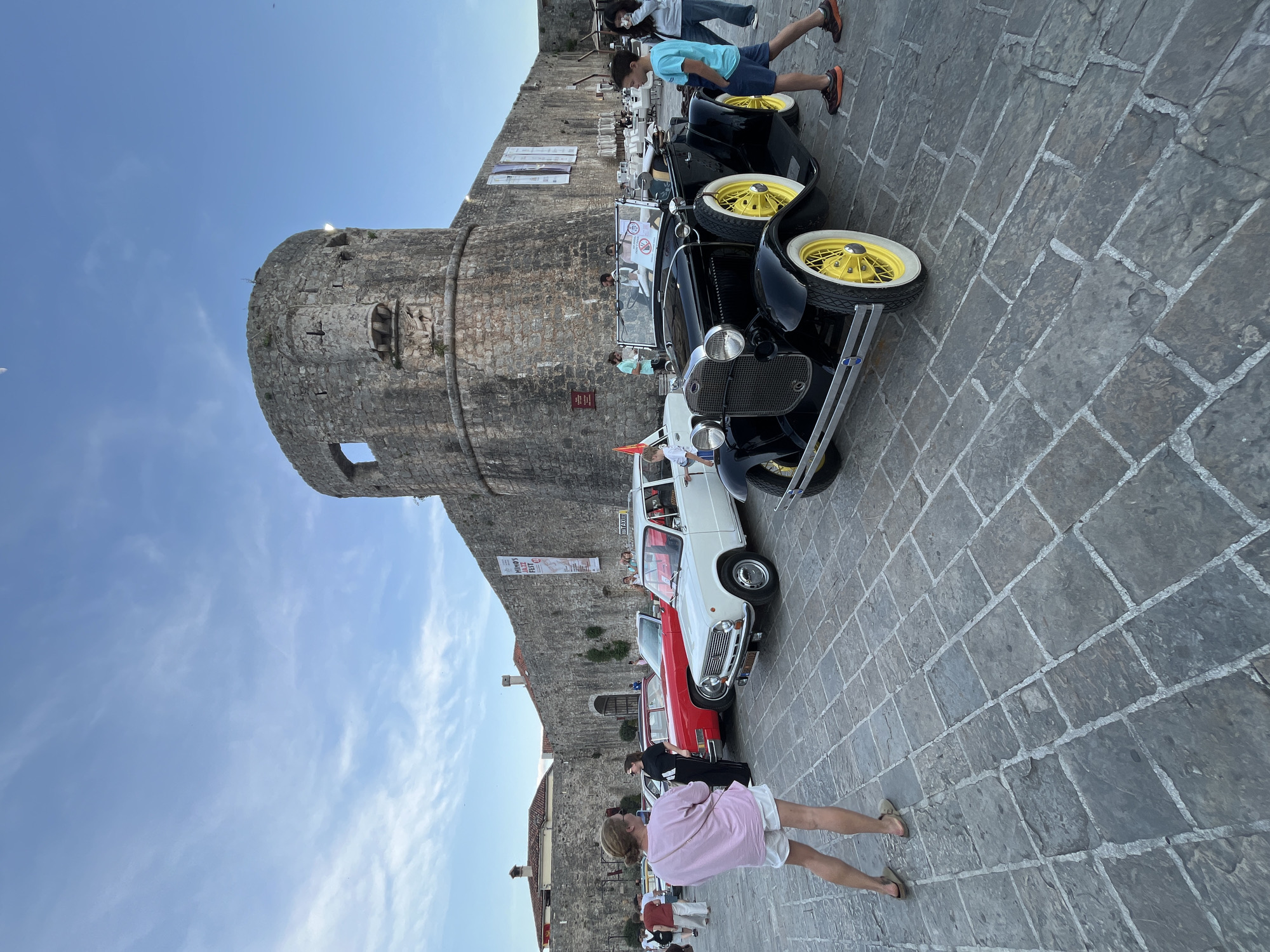 The image shows a scene in what appears to be a historic district. A medieval-looking stone tower is prominent on the left. Several vintage cars are parked along a stone-paved street. There are several people, with some inspecting the vehicles and others walking by. The sky is blue. The setting suggests a cultural or tourist area. I cannot confirm that this is the city of Budva, Montenegro.