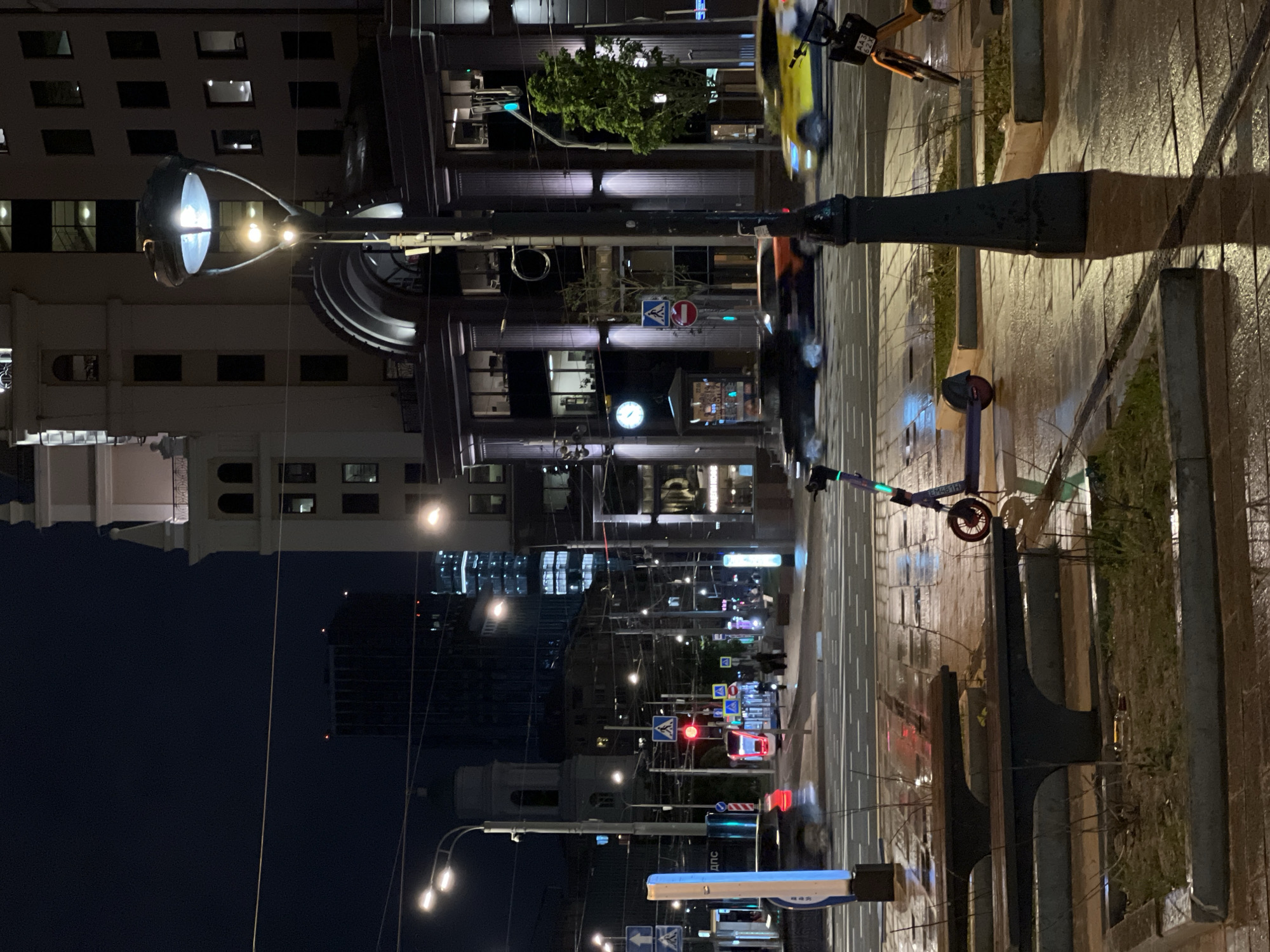 The image depicts a nighttime street scene in Moscow, Russia. The perspective is from street level, looking down a wet, paved road lined with buildings of varying heights and architectural styles. Streetlights illuminate the scene, reflecting off the wet pavement. A yellow taxi cab and electric scooters are parked or moving along the right side of the road. Traffic lights and pedestrian crossing signs are visible, indicating an urban environment. People can be vaguely seen walking in the distance. A clock is visible on one of the buildings. The overall atmosphere suggests it is an evening or night after rainfall.