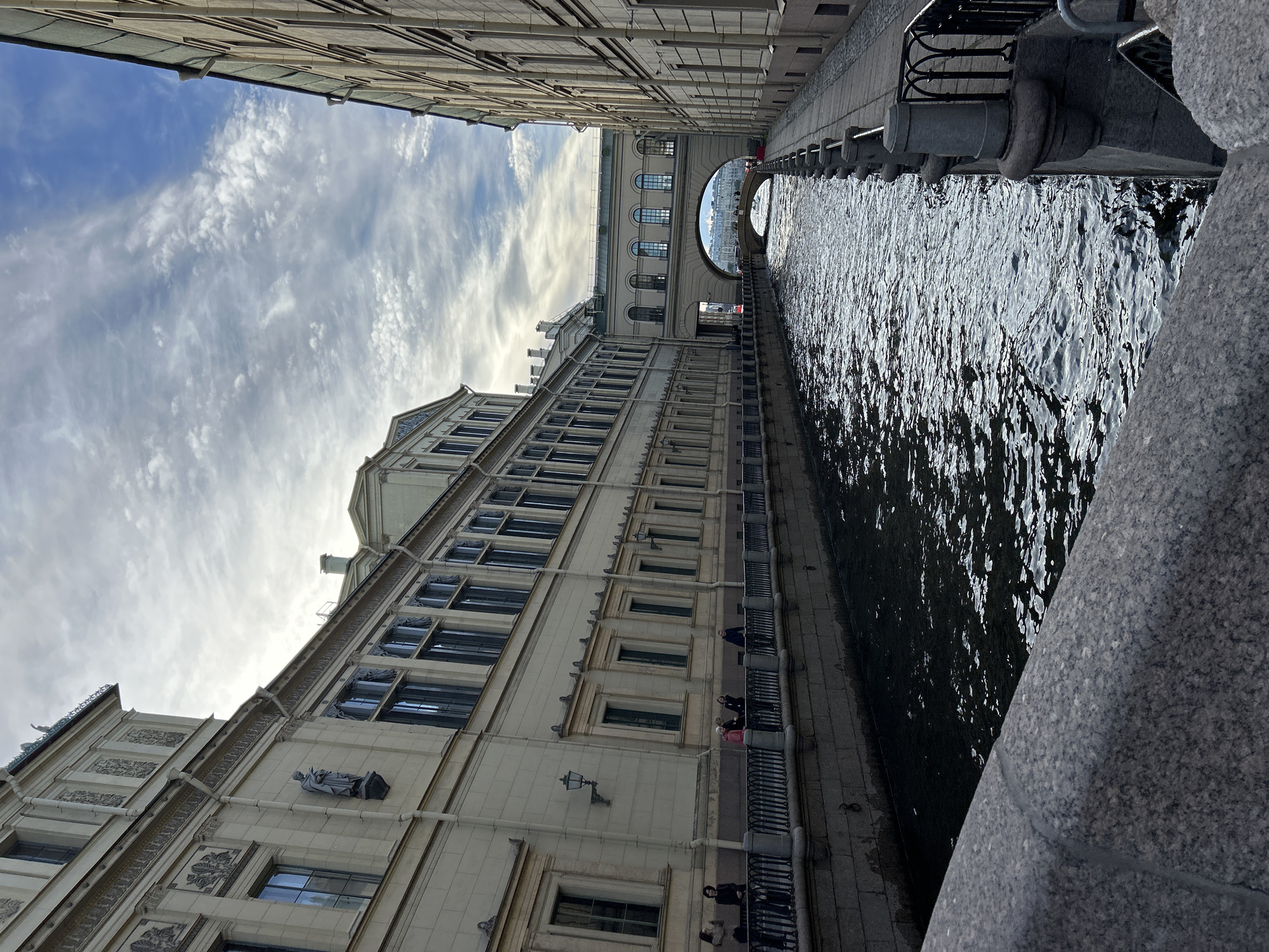 This media file captures a scenic view of a canal bordered by ornate buildings, with a bridge arching in the distance. The camera angle is tilted, giving a unique perspective of the architecture and water. Sunlight reflects off the rippling surface of the dark canal, creating a dynamic visual. Several people are visible on the elevated walkway along the canal, observing the surroundings or engaged in conversation. The buildings feature classical architectural elements, including numerous windows, decorative facades, and sculptures, suggesting a historical or European urban setting. The sky above is partly cloudy, with streaks of white clouds against a pale blue backdrop, indicating it could be daytime with a mix of sun and clouds. The overall mood is peaceful and contemplative, inviting viewers to appreciate the architectural beauty and the tranquil flow of the canal. No visible text is present in the image.