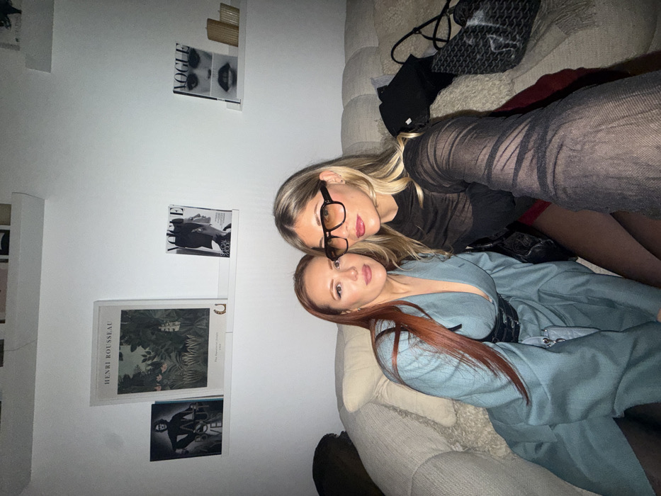 Two women pose on a beige sofa. The woman on top has blonde hair and wears glasses, a black top, and tights. Below her, the other woman has red hair and wears a light blue blazer dress with a black belt. A black bag is above them on the sofa. A white wall behind them has staggered shelves with magazines, posters, and small gold décor. One poster reads "HENRI ROUSSEAU".