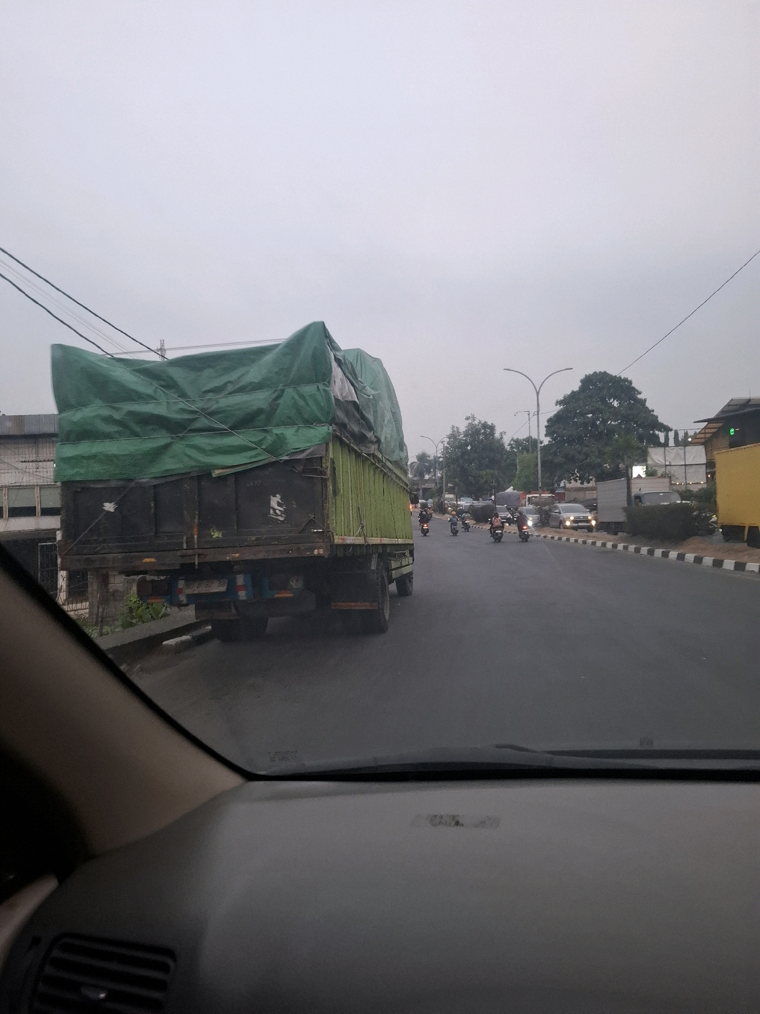 The image captures a view from inside a vehicle looking out onto a street in Citeureup, Indonesia. In the foreground, a portion of the vehicle's dashboard and windshield is visible. The main subject is a large, dark-colored truck with its cargo covered by a green tarp. The truck is in motion or stopped in traffic.
The street is paved and appears to be a main road with multiple lanes. There is moderate traffic, including several motorcycles with riders, cars, and at least one other truck in the distance. The environment suggests an urban or semi-urban setting with buildings visible on the left side of the road. Trees and streetlights are also present. The sky is overcast, indicating a cloudy or possibly dusk/dawn lighting condition. The overall impression is a typical street scene with vehicles navigating the road.