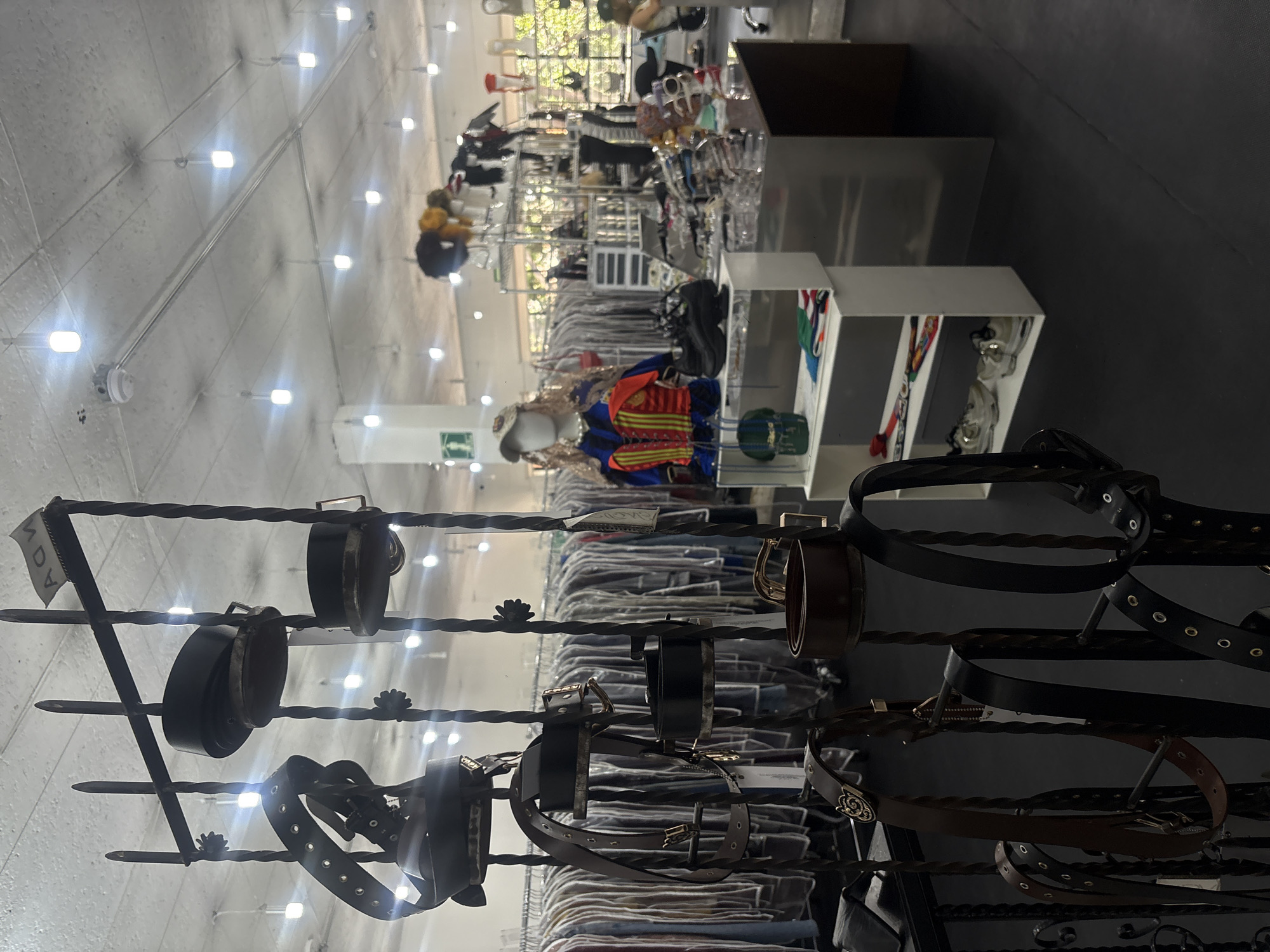 The image depicts the interior of a retail store, likely a clothing or accessories shop, in Mexico City, Mexico. The focus is on a wrought iron display rack holding various belts of different styles and colors. In the background, clothing racks are filled with garments. Further back, there's a display area with mannequins and shelving, suggesting the store offers a diverse range of fashion items. The store has bright overhead lighting. The overall impression is of a modern, well-lit retail space in an urban setting.