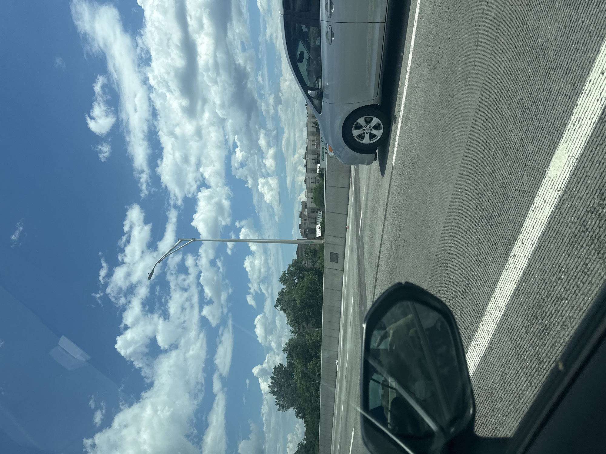The image captures an elevated highway scene in New York City on a bright, partly cloudy day, viewed from inside a moving vehicle. The upper portion of the frame features a clear blue sky interspersed with numerous white cumulus clouds. Faint reflections of overhead structures are visible on the windshield in the top left.
The roadway consists of a textured grey surface with parallel grooves and delineated by white lane markings. A light grey concrete barrier separates the highway from the adjacent landscape on the right.
A silver minivan, consistent with a Toyota Sienna model, travels in a lane to the right of the camera vehicle. Its front passenger side, including the wheel, side door, and a portion of the windshield, is visible. A faint impression of a driver can be discerned inside the minivan.
Beyond the concrete barrier, a dense canopy of green deciduous trees is visible, followed by multi-story, light-colored buildings in the background. One building exhibits a blue sign with white text that appears to read "477 BROADWAY," indicating the location is likely an elevated highway with a view towards Lower Manhattan or an adjacent borough. A grey street light pole with a curved arm is positioned to the right of the trees.
The lower left corner of the image shows the dark frame and side mirror of the vehicle from which the photograph was taken. The mirror reflects a distorted view, possibly of the vehicle's interior. No overt interactions between people or vehicles are depicted beyond typical highway travel.