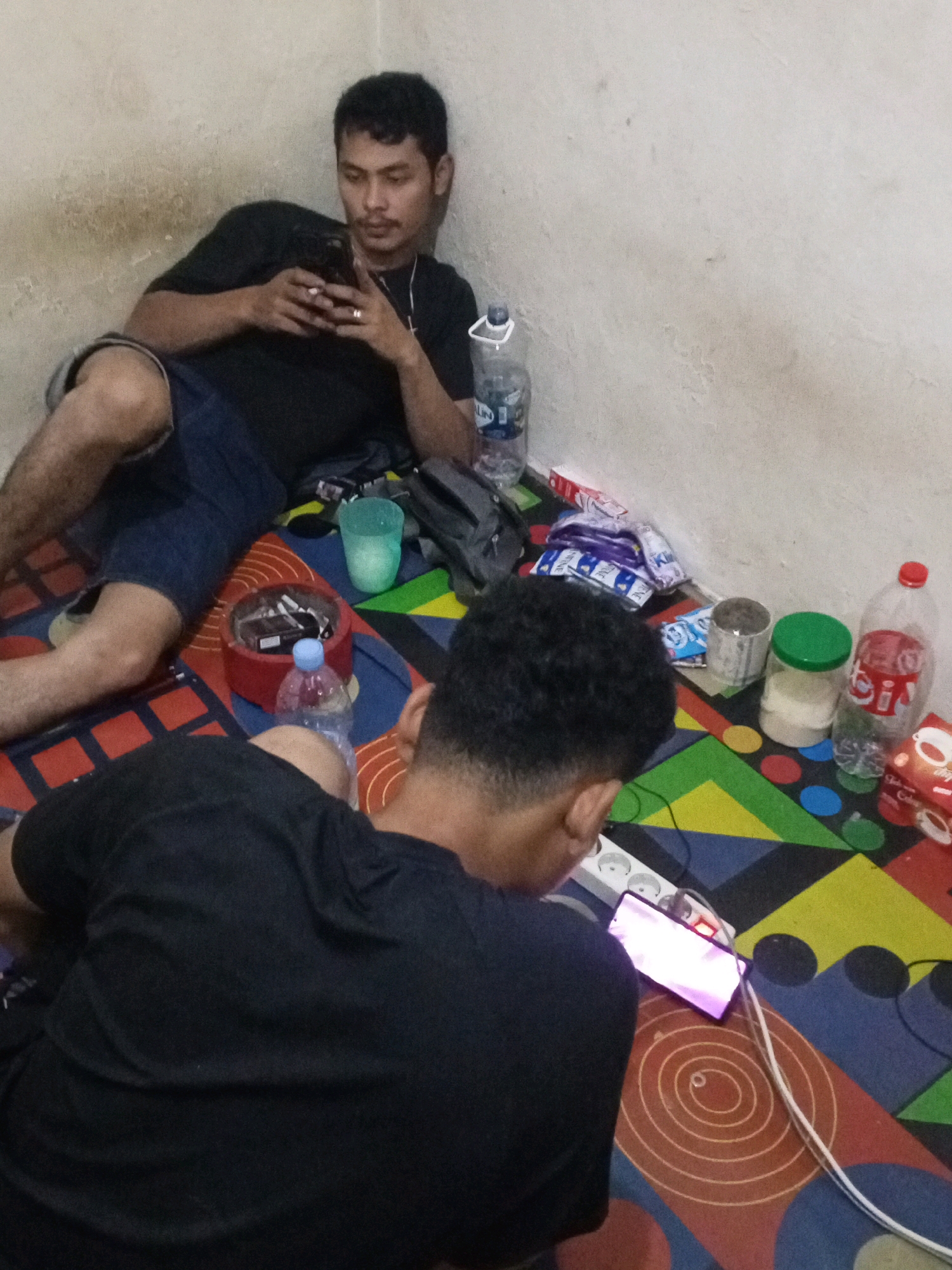 Two individuals are present in an indoor setting, likely a room, with a light-colored, textured wall forming the background and a colorful, patterned mat on the floor. The room appears to be in South Tangerang, Indonesia.
The individual on the left is leaning against the wall, lying on their side, and is wearing a black t-shirt and blue shorts. They are holding a mobile phone in their hands and looking at the screen. A lighter is visible in their left hand, suggesting they may have been smoking. Their right leg is bent, and their left leg is extended.
The individual on the right is lying face down on the floor, with their back to the camera. They are wearing a black t-shirt. A mobile phone is positioned in front of them on the floor, connected to a power strip via a white cable. The phone screen is illuminated.
Scattered around the floor are various objects: a nearly empty plastic water bottle with a blue cap, a glass with a light green liquid, a red ashtray filled with cigarette butts, several packets of snacks or cigarettes, two plastic jars (one with a green lid containing a white powder, the other an empty tin can), a large plastic bottle of liquid with a red cap, and a small box with a coffee logo. A power strip with multiple outlets is also visible on the floor. The overall impression is a casual, relaxed environment.