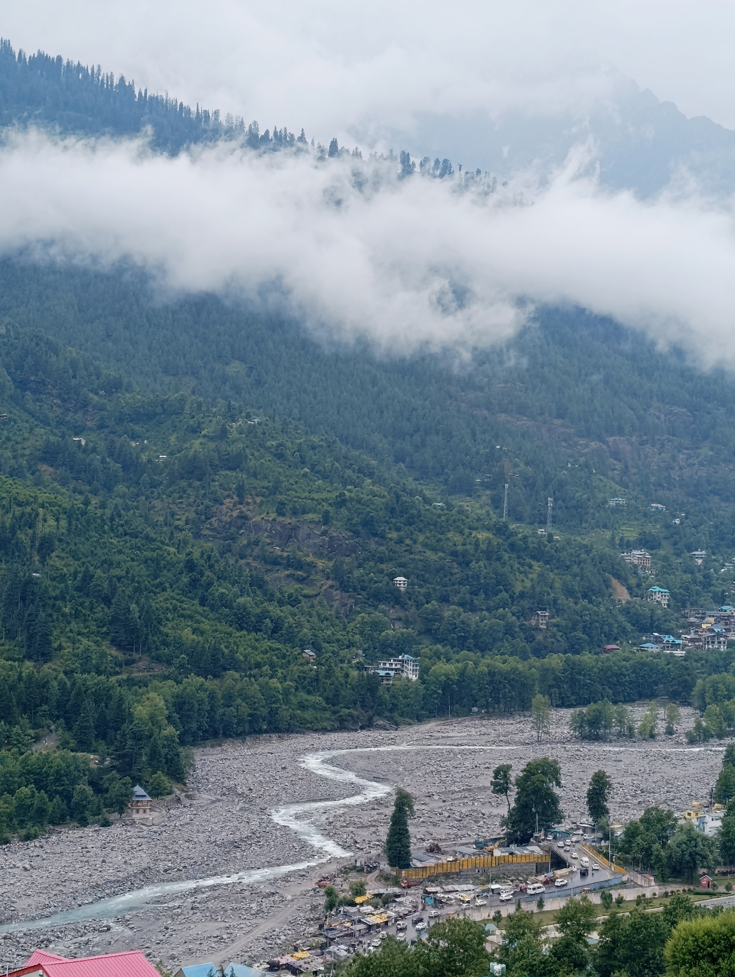 This image captures a scenic view of a mountain valley in Vashisht, India, likely during the monsoon season given the presence of clouds and fog. The scene is dominated by lush green mountainsides covered with dense pine trees. White clouds are drifting across the upper slopes, partially obscuring the mountain peaks and adding a dramatic effect.
In the mid-ground, a winding river flows through a wide, rocky riverbed, its water appearing a light blue or grey. Along the riverbanks and on the slopes, small clusters of buildings are visible, suggesting a settlement or town. Some of these structures appear to be houses, while others might be guesthouses or shops. There are also signs of infrastructure like telecommunication towers on the hills.
In the foreground, a bustling area with several vehicles, including cars and vans, is visible on a road. This area appears to be a marketplace or a hub of activity, with temporary stalls and shops set up. A distinctive structure, possibly a temple or a monument, with a pagoda-style roof, stands on the rocky terrain near the river. The overall impression is one of a natural landscape integrated with human habitation.