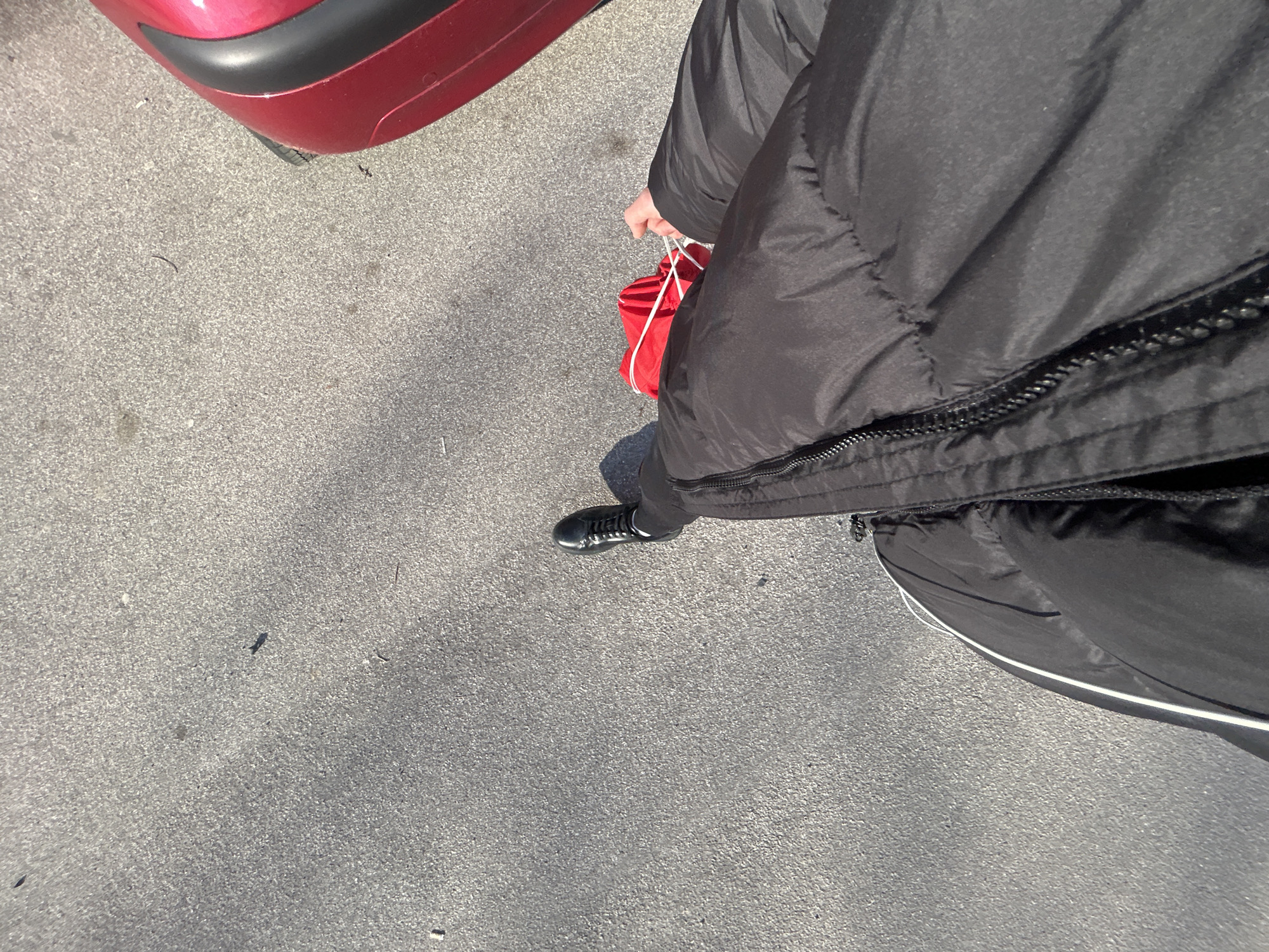 The image, shot from a high angle, shows a person standing on asphalt. The individual is wearing a black puffer jacket and black pants, with shiny black shoes. The person is holding a red bag with white drawstrings. A section of a red vehicle is visible in the upper left corner. The asphalt surface is gray and shows signs of wear. The specific location in Zlatograd, Bulgaria, cannot be determined from the image alone.