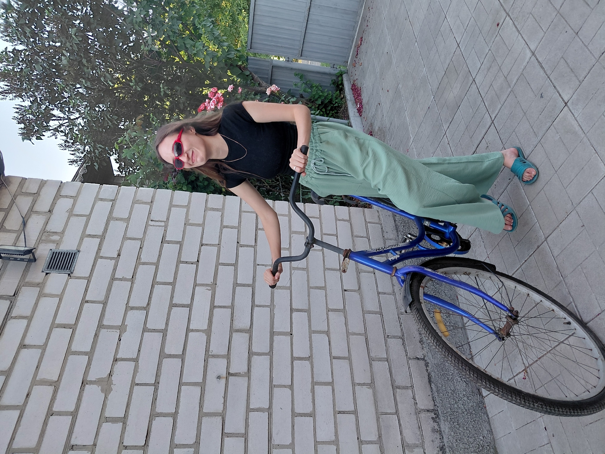 A young woman with long brown hair and red sunglasses is standing next to a blue bicycle on a paved walkway. She is wearing a black t-shirt and loose-fitting light green pants. She is holding onto the handlebars of the bicycle, which is positioned to her right. The bicycle has a blue frame, silver spokes, and a black tire. The paved walkway is made of gray square tiles. Behind the woman, there is a white brick wall and a green tree with pink flowers. The time of day appears to be daytime, and the weather seems pleasant. The scene is outdoors, possibly in a residential area or park. The woman's expression is neutral, and she appears to be posing for the picture. There is no visible text in the image.