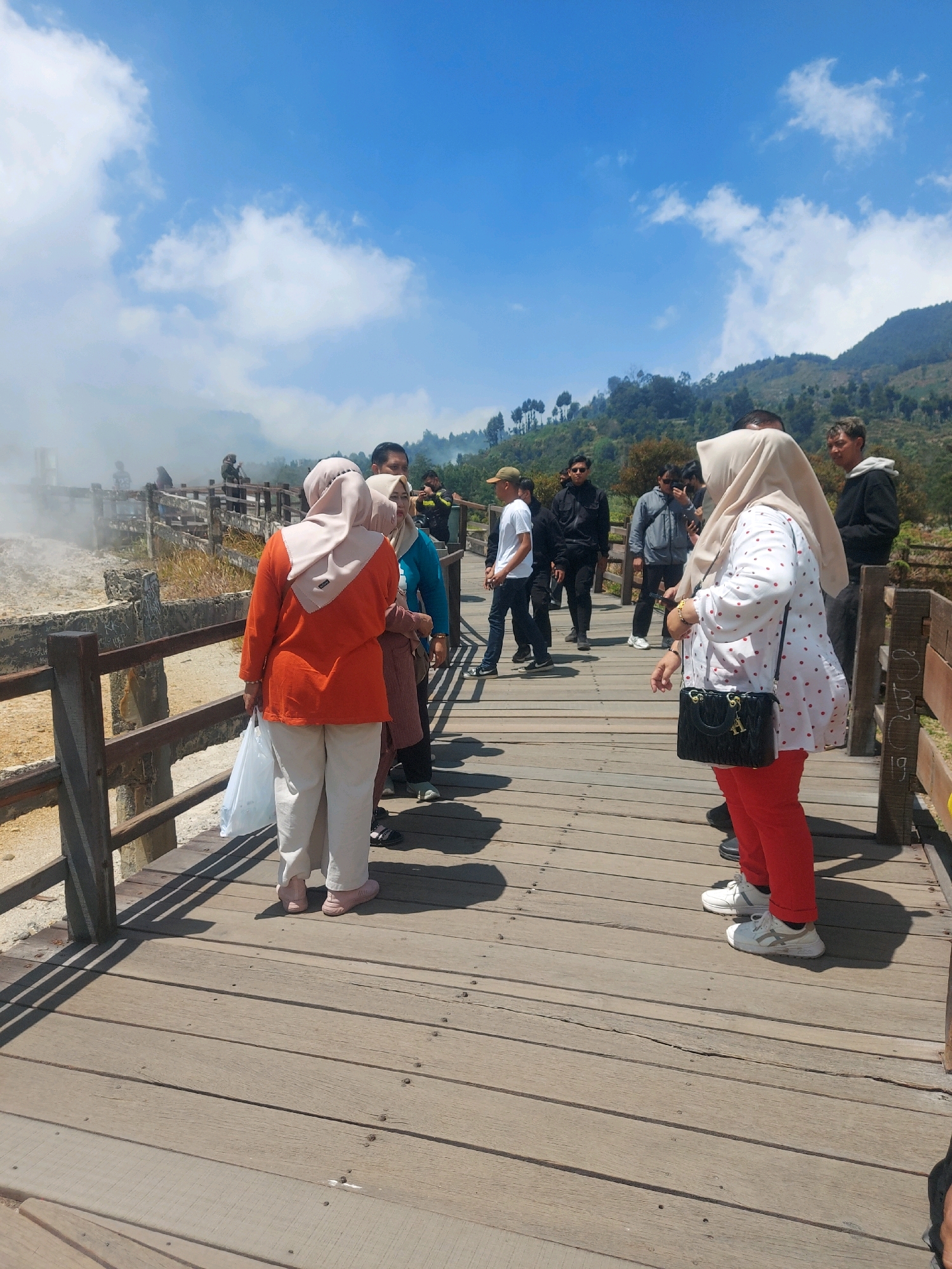 The image shows a wooden boardwalk at the Sikunang crater in Indonesia, a geothermal area known for its sulfurous fumes. Several individuals are visible on the boardwalk. In the foreground on the left, a woman wearing an orange shirt and light-colored pants, with a light-colored headscarf, is holding a white plastic bag. To her right, another woman in a brown top and headscarf is partially visible. Further ahead, a woman in a white polka-dot shirt and red pants, wearing a light-colored headscarf and carrying a black purse, stands with her back mostly to the camera. On the right side of the boardwalk, a person in a black hoodie and red pants is partially visible, wearing white sneakers. Several other people, mostly men, are scattered further down the boardwalk. The background features the crater's steamy landscape on the left, with lush green hills and a bright blue sky with white clouds above. The boardwalk is constructed of weathered wooden planks and has wooden railings.