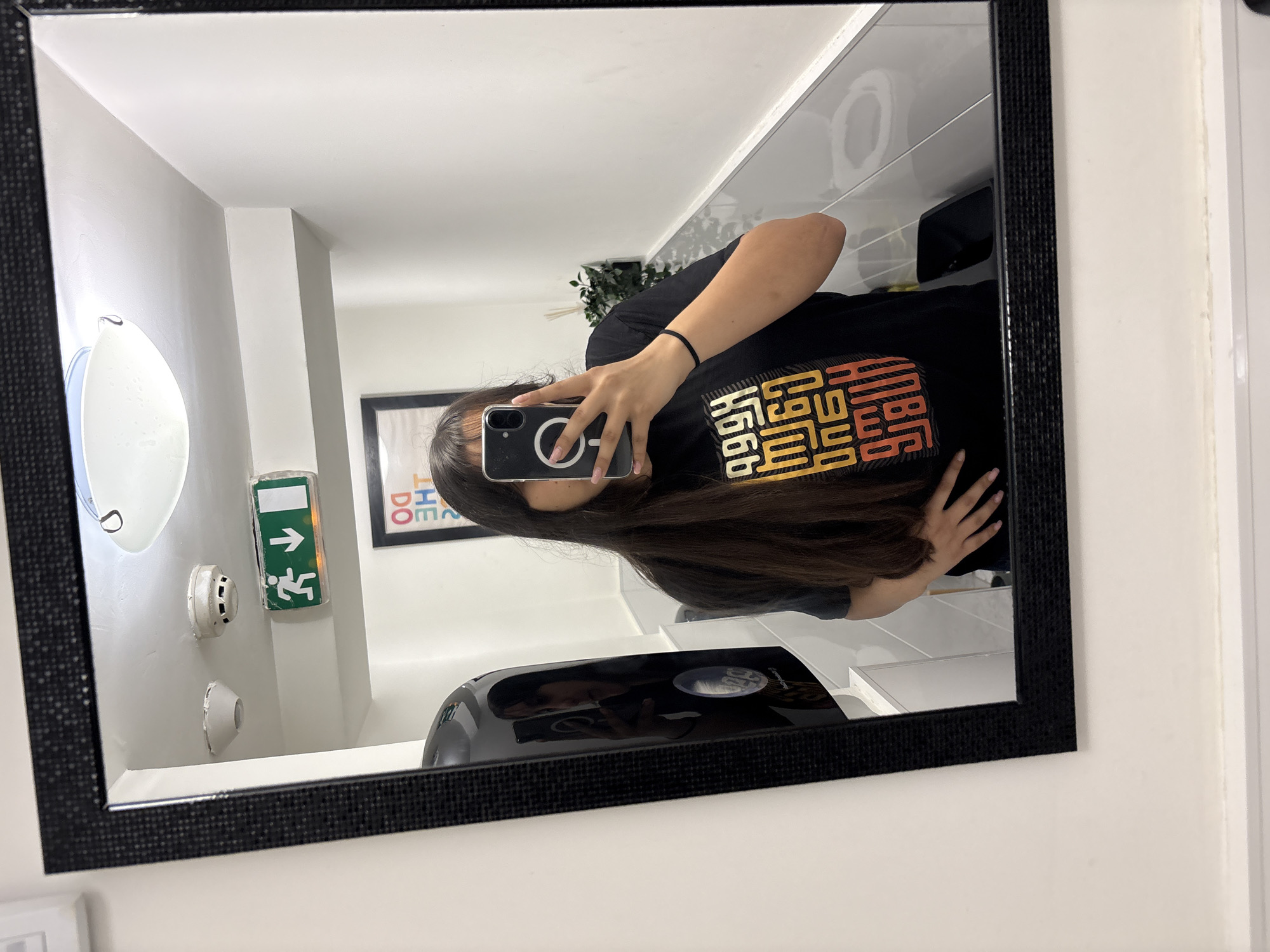 A person is holding a phone and taking a picture of themselves in a mirror. The mirror is mounted on a white wall, and the reflection shows a bathroom interior, including a sink, soap dispenser, light fixture, exit sign, and a framed poster. The person is wearing a black t-shirt. The specific location cannot be identified from the image.
