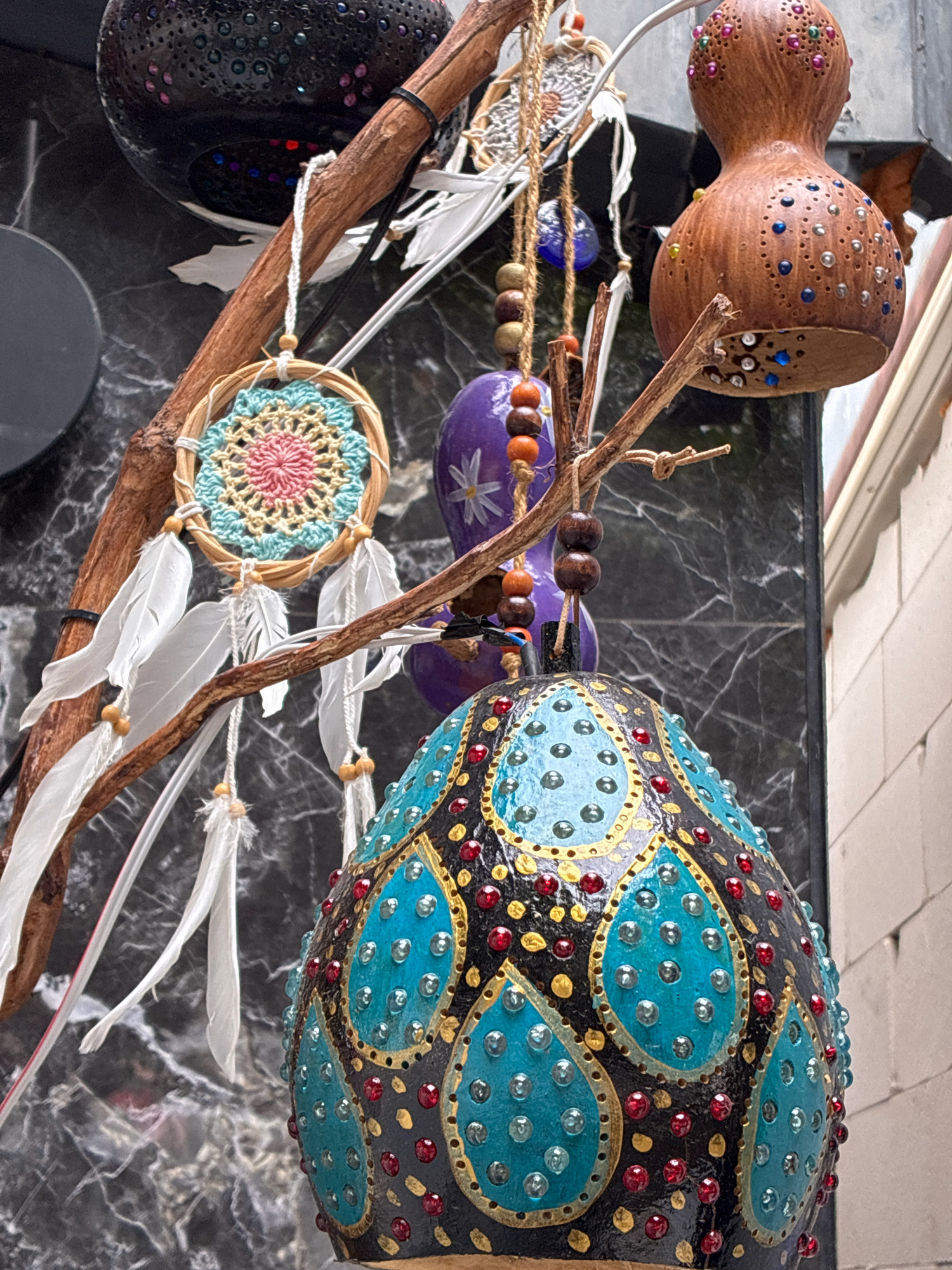 The image captures a close-up view of various handcrafted decorative items hanging from branches, possibly for sale in a market or shop in Alanya, Turkey. In the foreground, a large, ovate object, likely a decorated gourd or ceramic, is prominent. It's painted black with intricate patterns in teal and gold, studded with small red and clear rhinestones or beads. To the left, a small dreamcatcher with a crocheted center in pink, yellow, and blue, adorned with white feathers, hangs from a branch. Further up and to the left, a black, perforated lantern-like object with colorful accents is visible. To the right, a light brown, gourd-shaped object, also decorated with colorful beads, is suspended. Between these items, purple, egg-shaped decorations with white floral designs and strings of wooden beads are also present. The background consists of a dark, marbled surface and a lighter, textured wall, suggesting an outdoor or semi-outdoor setting. The overall impression is one of artisanal crafts, possibly reflecting local artistry and tourism in Alanya.