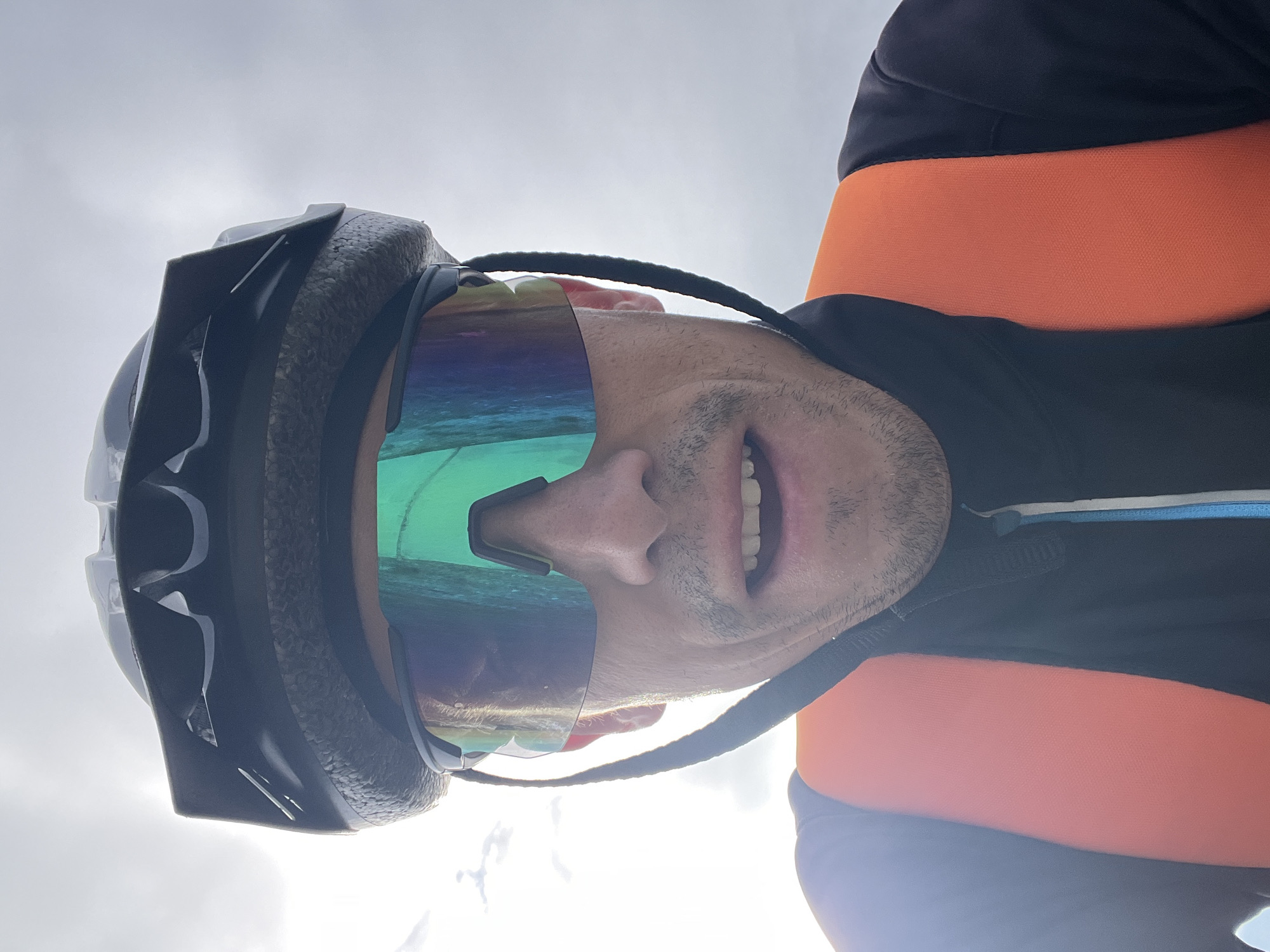 A close-up, eye-level, selfie shot shows a man wearing a black bicycle helmet and large, reflective, green and blue tinted sports sunglasses. He is wearing a black jacket with a high collar and an orange strap across his chest, possibly part of a backpack or safety vest. The man is looking directly at the camera with his mouth slightly open, revealing teeth. His facial hair includes a mustache and a light stubble on his chin. The background is a bright, overcast sky with some indistinct cloud formations. No specific location in Chaikovsky, Russia, can be identified from the image.