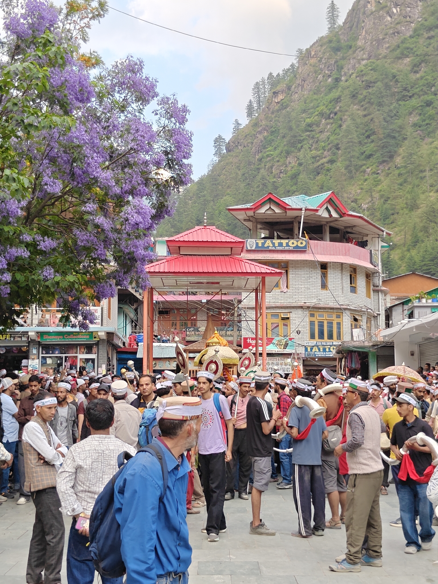 A bustling street scene in Manikaran, India, captures a vibrant cultural gathering under a bright daytime sky. A large crowd of men, many wearing distinct white Himachali caps with colorful bands, fills the foreground and midground, suggesting a procession or festival. Some individuals carry ornate golden structures, possibly palanquins or temple chariots, while others hold large, curved traditional musical horns, indicative of a religious or cultural event.
To the left, a magnificent tree with abundant purple blossoms extends over the scene, adding a splash of color. Behind the crowd, multi-story buildings with distinctive Himachali architecture, featuring colorful red and green roofs, line the street. A prominent building advertises "TATTOO" and "SENIK INK ZONE" with a mobile number "90827-00725." Other visible shop signs include "Shanaya Collections," "Cafe BLUE WOR," and "HAA SWEETS & FAST FC COME" (likely 'HOME' or 'CORNER'). A phone number "6230067166" is also visible. In the background, a steep, forested mountain rises majestically, characteristic of the Himalayan landscape. The atmosphere appears lively and communal.
