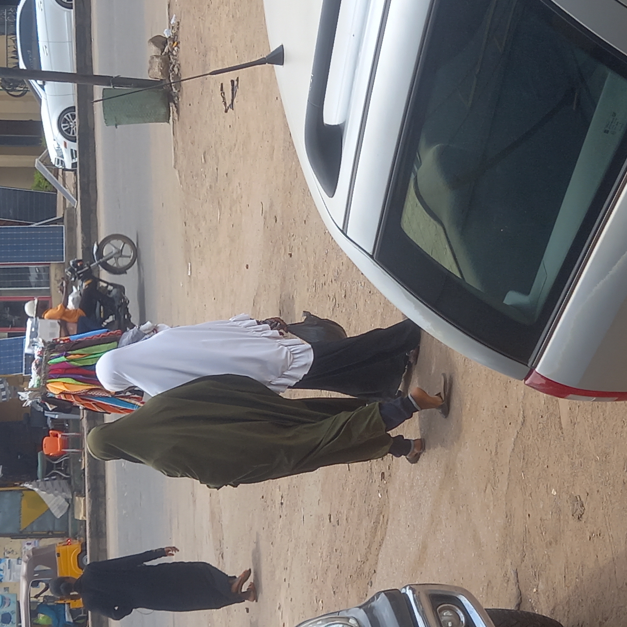 The image captures a street scene in Dadi Riba, Nigeria. A white car dominates the right side of the frame. Three women are walking along the street: one wearing a green hijab, another in a white hijab, and the third in a black garment. In the background, a motorcycle is parked, and colorful clothing items are displayed for sale. Solar panels are visible on a nearby building. The ground appears to be a dirt road.