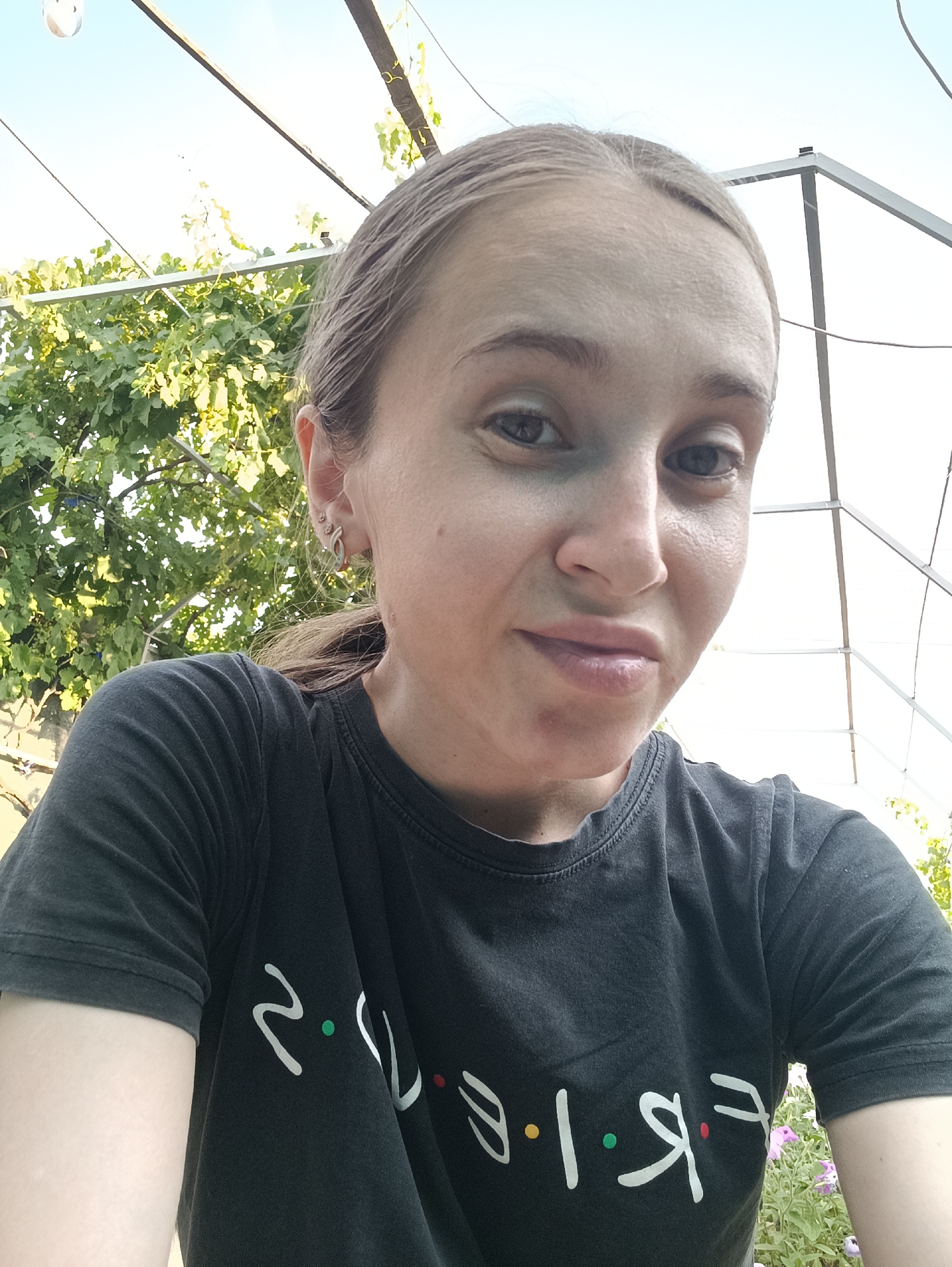 A selfie of a young woman with blonde hair pulled back in a ponytail, wearing a dark t-shirt with the word "Friends" printed on it in white lettering. She has earrings in her left ear and appears to be outdoors, with greenery and a bright sky in the background. The location is Vypasne, Ukraine.