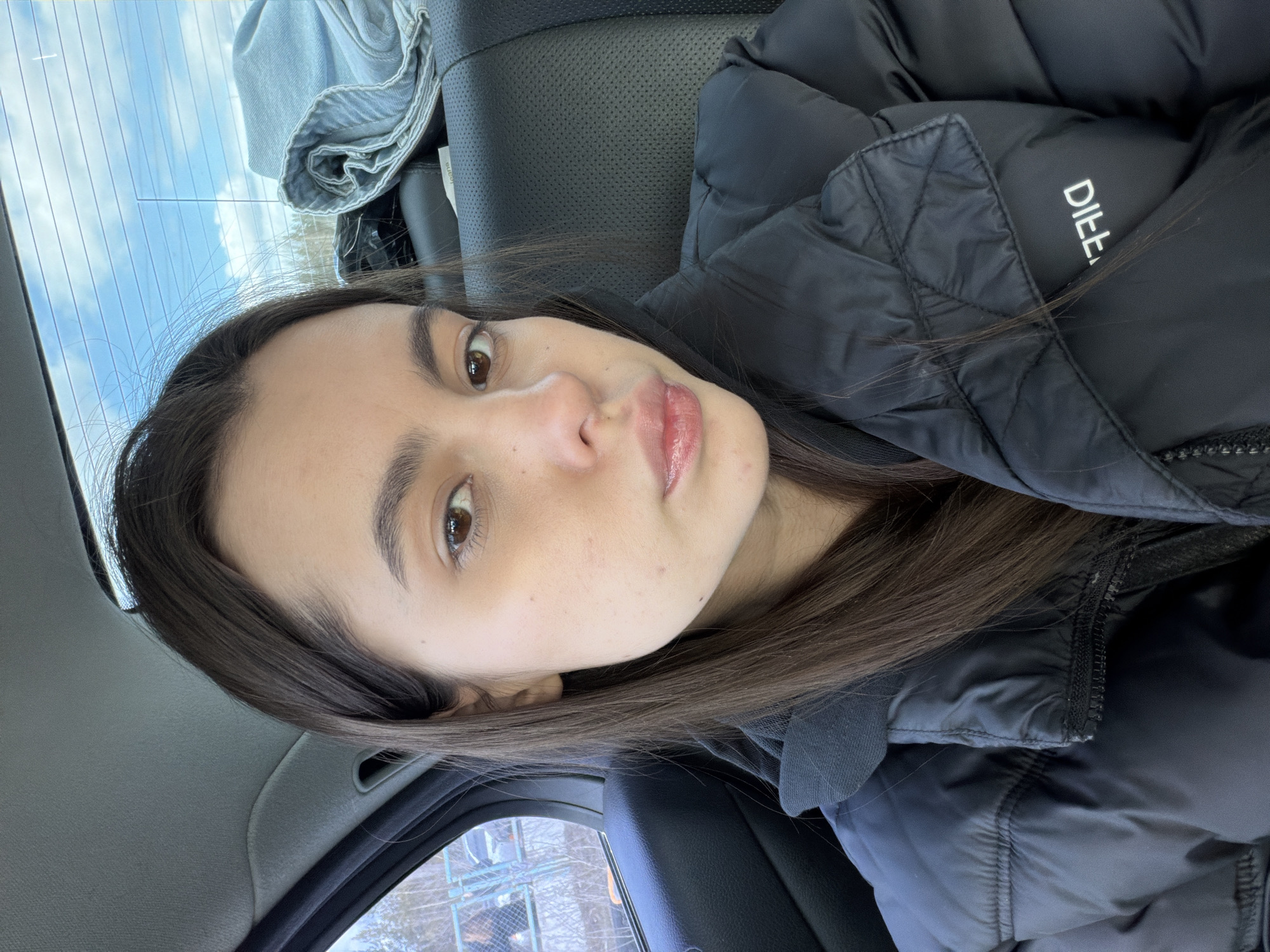 A young woman with brown eyes and hair is sitting in a car, likely in the backseat, wearing a black puffer jacket. The car interior is gray and black. Outside the car window, a glimpse of a snowy landscape with a fence and buildings is visible. The photo might be taken in Perm, Russia as the model's ethnicity can be traced back there.