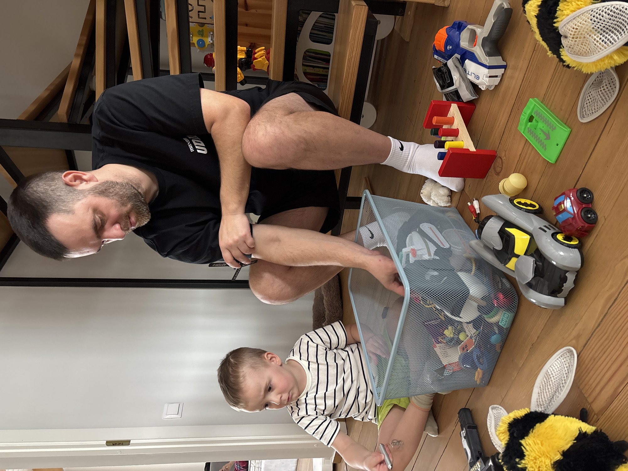 A man and a child are looking into a blue mesh laundry basket on a wooden floor. The man has a beard and is wearing a black t-shirt and black shorts with white socks. The child is wearing a striped shirt and is kneeling on the floor. The laundry basket is filled with various toys. Other toys are scattered around the basket, including a toy gun, a wooden toy, and a toy car. There are wooden stairs in the background. The location is not identifiable as the city of Chaikovsky, Russia, from the given visual information.
