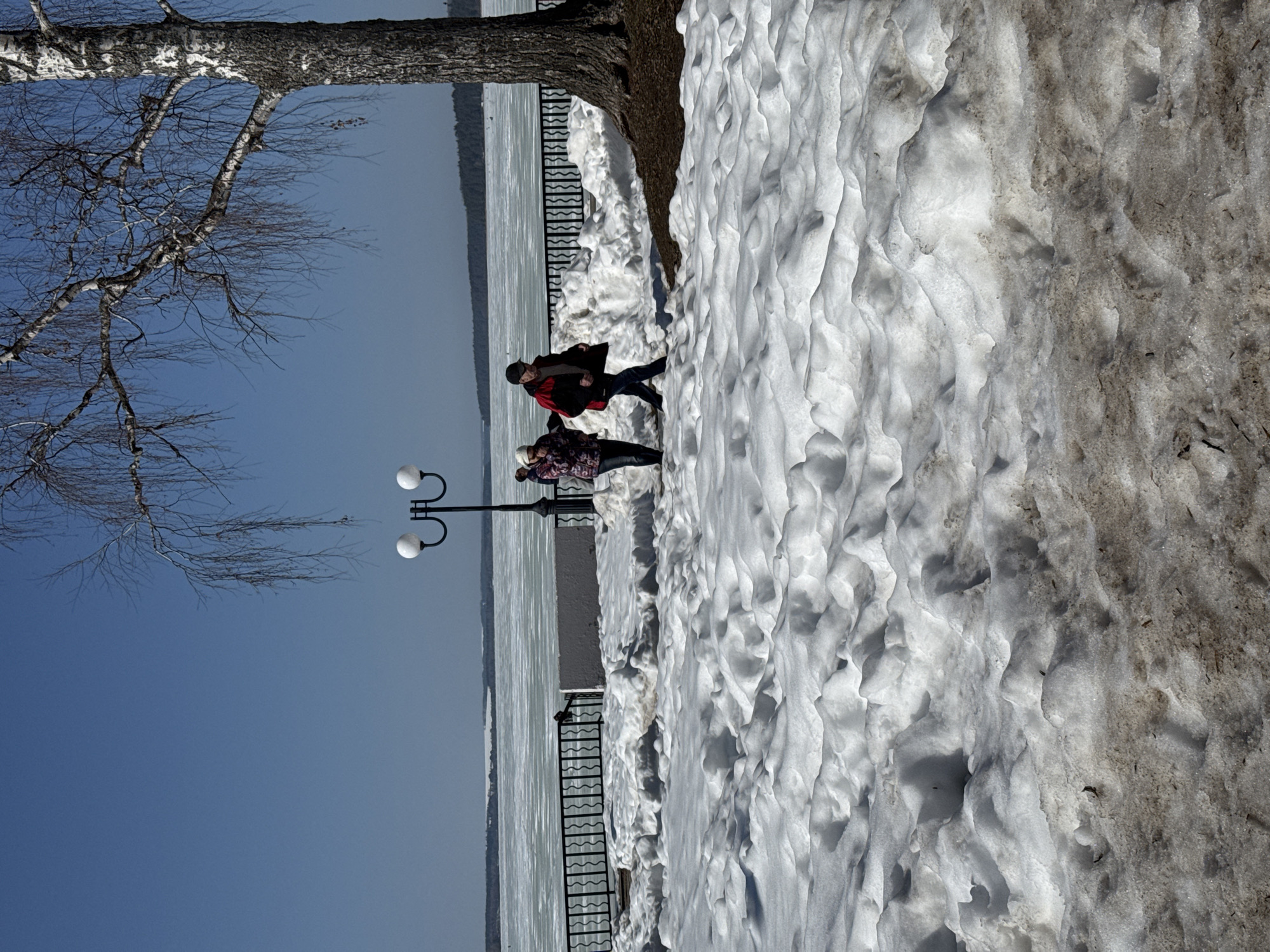 The image depicts a winter scene along the embankment of the Votkinsk reservoir in the city of Votkinsk, Russia. Two people are walking along a snow-covered path beside a black metal fence. The frozen surface of the reservoir stretches out to the horizon under a clear blue sky. A tree with bare branches is visible on the left side of the image, and a street lamp stands near the walking figures. A large snowdrift lines the path.