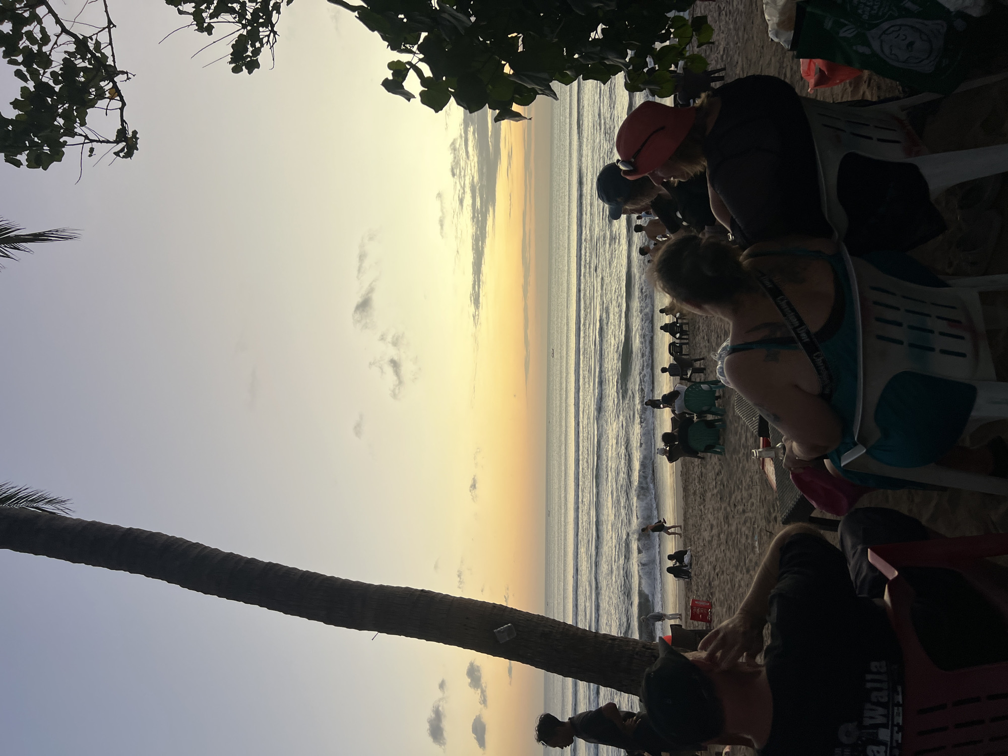 The image captures a serene beach scene in Kuta, Indonesia, during what appears to be sunset or dusk. The sky is a gradient of soft blues and oranges, with scattered clouds, and the ocean gently laps the shore. In the foreground, several people are seated in chairs on the sand, facing the sea. A large palm tree trunk stands prominently on the left side of the frame, its fronds silhouetted against the sky. To the right, lush green foliage frames the upper right corner of the image.
On the beach, groups of people are visible, some standing and others sitting. Waves are breaking on the shore, and a few small boats are dotted on the horizon. The atmosphere seems relaxed, with individuals and small groups enjoying the view. The composition suggests a popular spot for evening relaxation, likely a restaurant or cafe with seating arrangements on the beach. The lighting indicates the transition from day to night, creating a warm and tranquil ambiance.