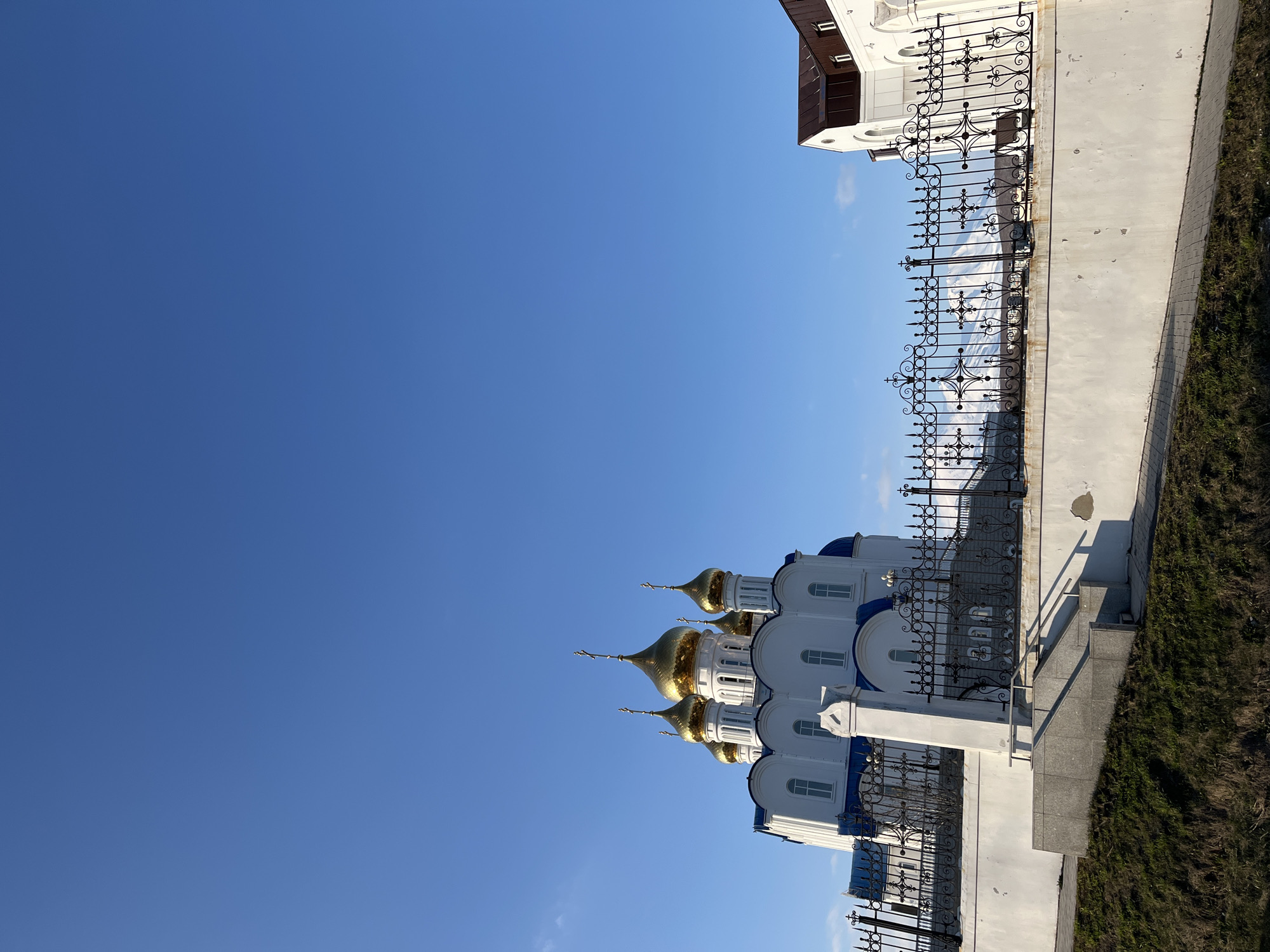 The image is an upward-tilted shot of the Holy Trinity Cathedral in Petropavlovsk-Kamchatsky, Russia. The cathedral's architecture includes white walls, blue domes, and golden cupolas, typical of Russian Orthodox churches. An ornate black metal fence lines a portion of the cathedral grounds. The foreground includes a concrete walkway and a grassy embankment. The sky is clear blue, with no visible clouds. No people are visible in the image.