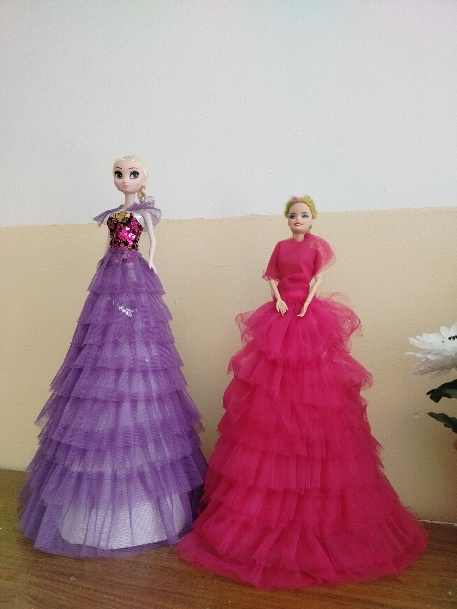 Two dolls are standing on a wooden surface against a light-colored wall. The doll on the left has blonde hair and is wearing a sequined pink bodice and a multi-layered purple tulle gown. The doll on the right has blonde and pink hair and is wearing a pink top with short sleeves and a matching multi-layered pink tulle gown. A white artificial flower with green leaves is partially visible in the lower right corner of the image. The scene is indoors and appears to be a domestic setting. There is no indication of the specific location within the city of Көлкент, Kazakhstan.