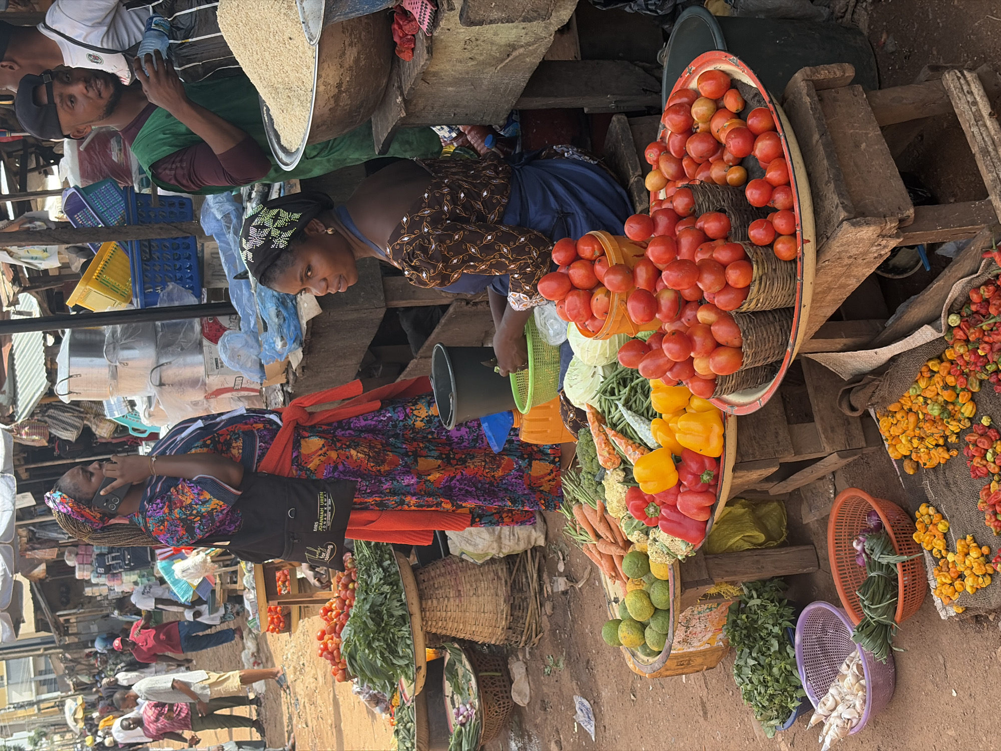 The image depicts a busy outdoor market scene in Jos, Nigeria. In the foreground, a vendor, a woman with dark skin wearing a blue apron over a brown and white patterned top, sits behind a display of fresh produce. Various vegetables like tomatoes, peppers, carrots, cauliflower, and green beans are neatly arranged on wooden tables and in woven baskets. To her left, a large bowl overflows with a mound of white rice, and a man wearing a green vest and a black cap stands nearby, holding a water bottle.
Further back, a woman in a colorful, patterned dress is talking on her phone, a black bag slung over her shoulder. Other market-goers, dressed in casual attire, navigate the crowded aisles. Stalls with awnings and various goods are visible in the background, suggesting a vibrant commercial atmosphere. The ground is a mix of dirt and sand, typical of an open-air market. The overall impression is one of a lively and bustling marketplace providing fresh produce and other goods to the community.