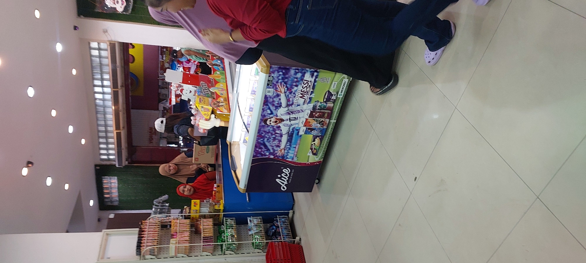 The image depicts an indoor scene, likely a convenience store or a small supermarket, with a tiled floor. In the foreground, a woman wearing a purple hijab and a red long-sleeved shirt is partially visible, with her arms raised as if gesturing. To her right, another person in dark blue jeans and a red long-sleeved shirt is also visible from the knees down, wearing light purple crocs.
Further into the scene, a large freezer chest is prominent, adorned with an advertisement featuring Lionel Messi in an Argentinian football jersey. The freezer is filled with ice cream products, some of which are visible on top. Next to the freezer, a blue counter is present, where two women are interacting. One woman, wearing a brown hijab, is facing away from the camera, and next to her, a woman in a red hijab is looking towards the counter. Behind the counter, shelves stocked with various food and drink items are visible.
To the left of the counter, a tall white shelving unit is filled with packaged goods, and a red shopping basket is placed on the floor. In the background, there are more shelves and displays, with a partially visible sign that appears to say "YOHAN". The ceiling features multiple light fixtures. The overall impression is a retail environment with customers and staff present. This scene is consistent with a convenience store in the city of Jayapura, Indonesia, given the typical appearance and merchandise seen in such establishments in that region.