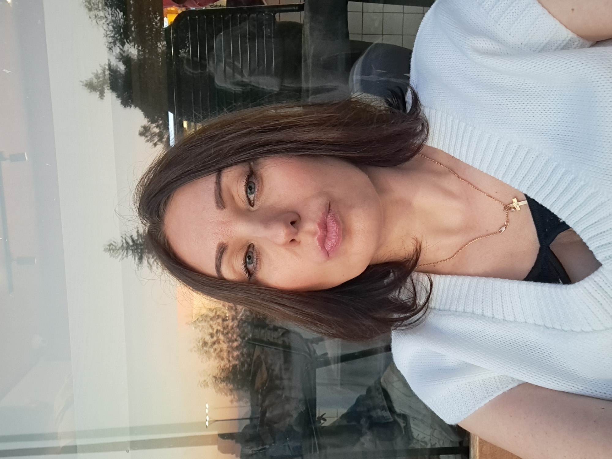 A mid-shot selfie of a woman with dark brown hair and fair skin. She is wearing a white knitted vest over a black garment, and a thin gold necklace with a cross pendant. Her lips are pursed. She is indoors, with a window behind her that reflects an outdoor scene at dusk. The outdoor scene shows trees, streetlights, and what appear to be buildings and possibly people. The image is likely taken in Irkutsk, Russia, given the context.