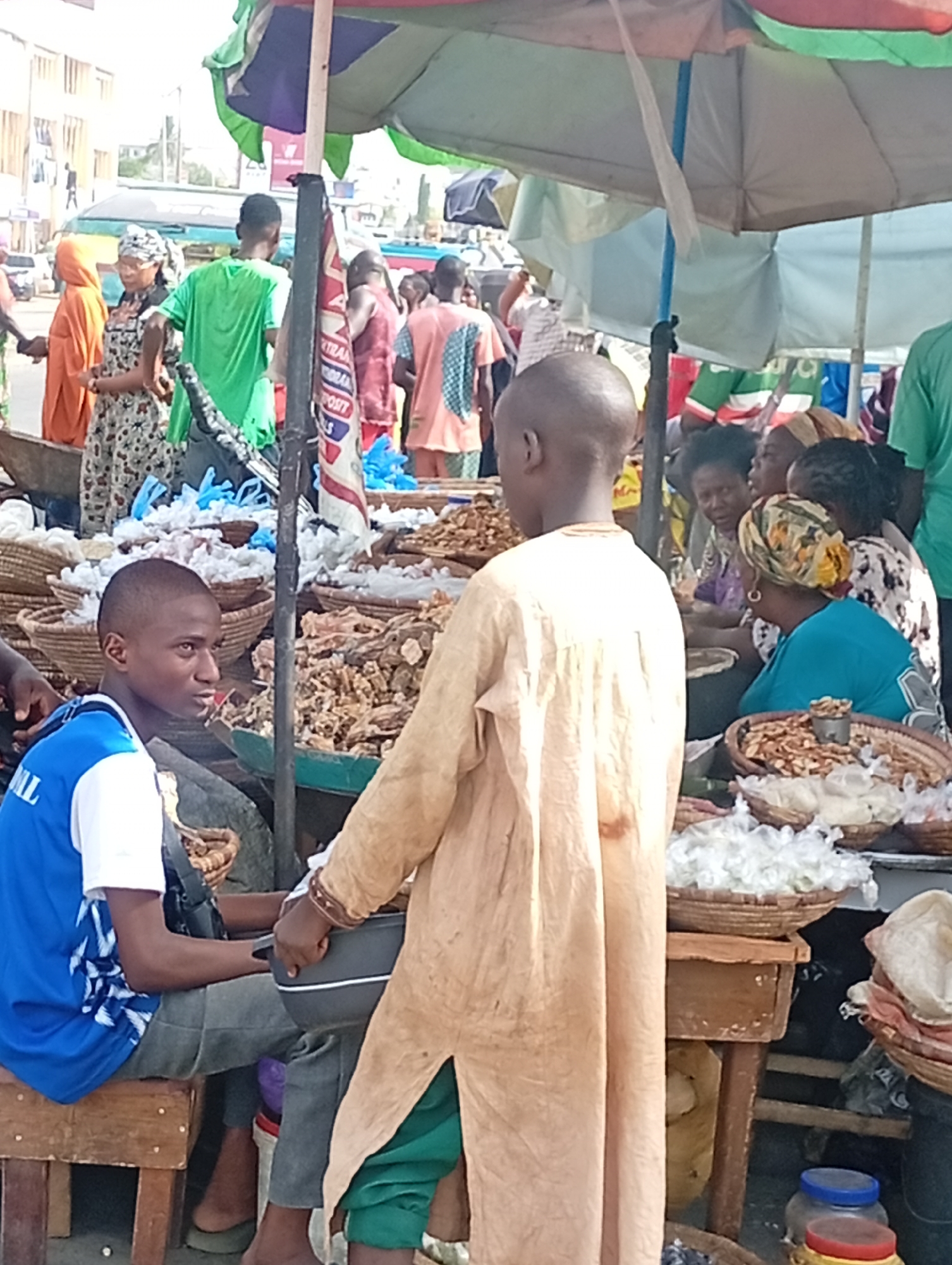 The image depicts a bustling outdoor market scene in Maiburiji, Nigeria. Numerous people are present, including vendors and customers. Items, likely foodstuff, are displayed in baskets on tables. A boy in a blue and white shirt sits on a wooden stool while another boy in a light-colored tunic holds a dark container. Women are seated behind the tables, presumably selling goods. An umbrella provides shade, and buildings are visible in the background. The overall impression is one of a vibrant and active trading environment.
