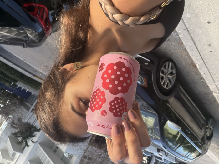 The image displays a close-up of an individual partially obscuring their face with a pink beverage can, set against an urban background in Miami, Florida, United States.
**People:**
A person is visible from the chest up, with only the right side of their face, including their right eye, eyebrow, and ear, exposed. They possess brown hair. A light-colored stud earring with a "Miu Miu" logo is present in their right ear. Their right hand holds the can, with fingernails coated in light pink polish. They are wearing a black top or dress, and a braided light-beige strap, likely from a bag or purse, rests on their left shoulder.
**Subjects/Objects:**
The central object is a cylindrical pink beverage can, held upright. The can features red, polka-dotted berry or flower-like designs and legible text including "Strawberry Rose," "Premium Sparkling Water," "5g Sugar," and "Soda's back, but Better."
In the background, a dark-colored Volkswagen Golf Alltrack station wagon with silver side mirrors and multi-spoke alloy wheels is parked parallel to a concrete curb. Further down the street, additional light-colored sedans are visible. The rear portion of another dark-colored vehicle is partially visible on the far right.
**Environment:**
The scene is outdoors in bright daylight. A concrete sidewalk with a discernible curb transitions to an asphalt road. On the right, a multi-story white building with numerous balconies rises diagonally into the background, characteristic of modern Miami architecture. Palm trees and other sparse vegetation are visible between the street and the building. A clear sky is discernible in the upper right quadrant.
