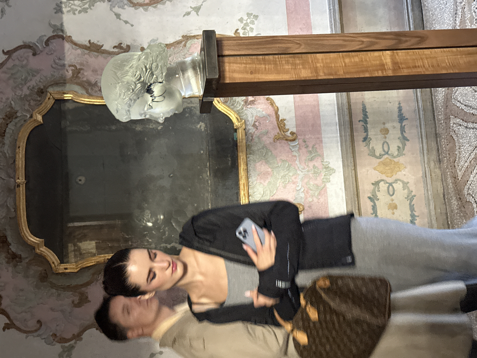 A young couple is reflected in an ornate mirror in Venice, Italy. The woman holds a phone, possibly taking a photo. She wears a grey tank top, a black jacket, and grey pants. A Louis Vuitton handbag sits beside her. The man wears a tan jacket. The ornate mirror is affixed to a wall decorated with floral patterns. A glass sculpture of a head sits atop the mirror. The room is decorated with vertical stripes of pink, white, and gold.