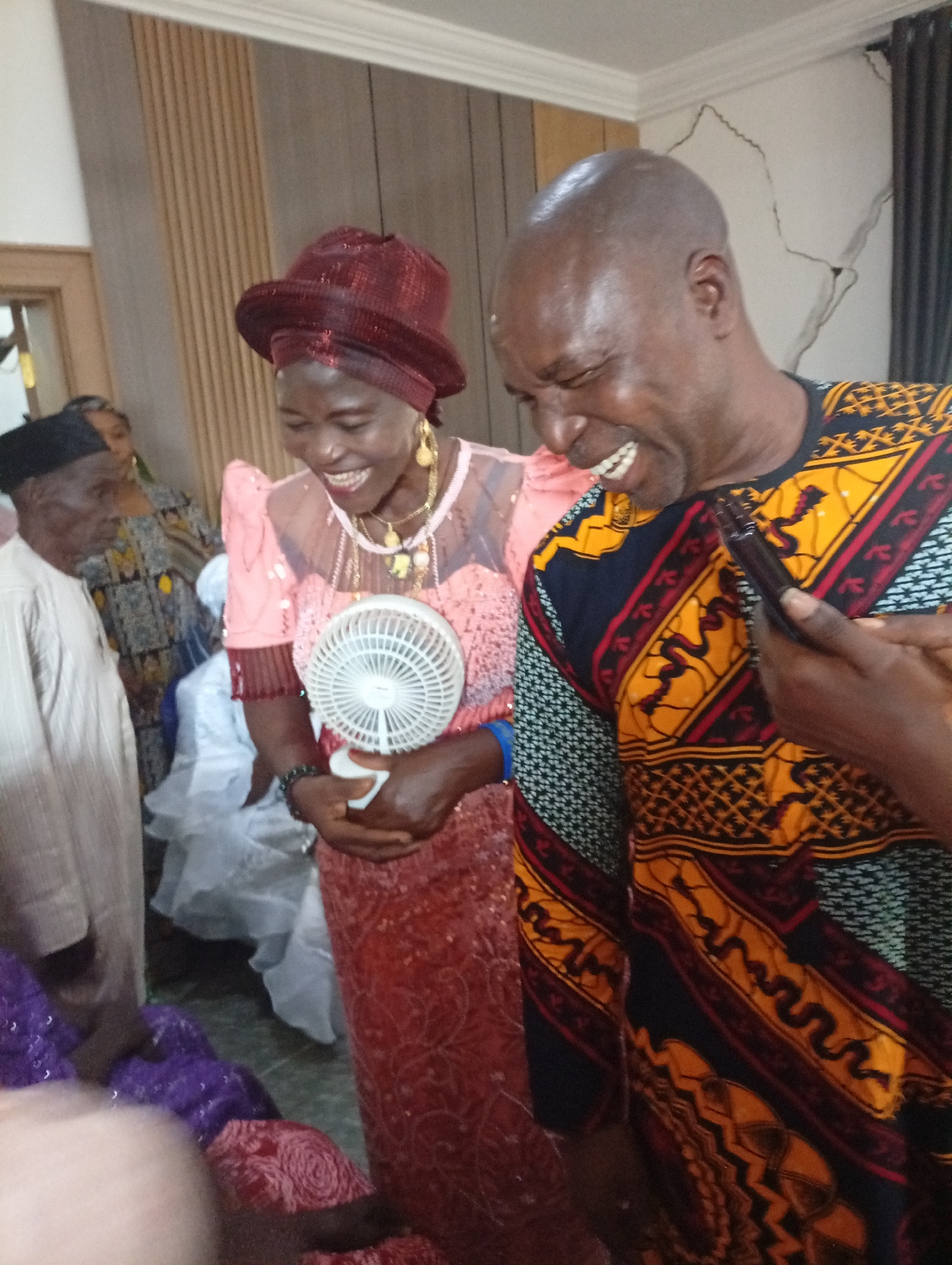 A medium shot captures a festive scene in Sabon Tasha, Nigeria, where a man and woman are laughing heartily. The woman, adorned in a vibrant pink dress and a red headscarf, holds a small white fan. The man, dressed in a colorful patterned shirt, is looking at his phone. Other individuals, some in traditional attire, are visible in the background, suggesting a gathering or celebration. The setting appears to be indoors, with wooden paneled walls and an off-white ceiling. The overall atmosphere is joyful and engaging.