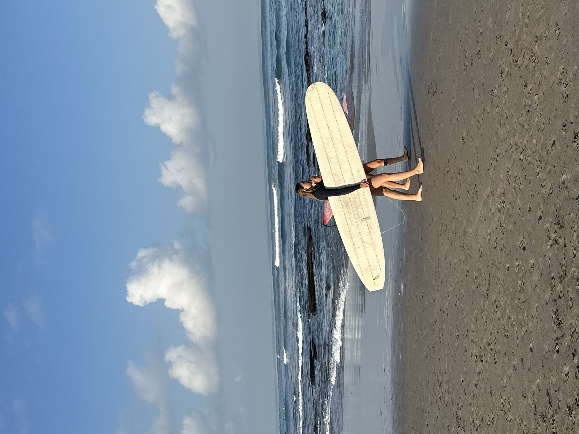 Two individuals, likely women, are walking along a sandy beach in North Kuta, Indonesia, carrying surfboards. The individual in the foreground is wearing a black wetsuit and carrying a light-colored surfboard horizontally. The second individual, partially obscured, is behind and to the right of the first, carrying a pink surfboard. They are walking on wet sand near the shoreline, with gentle waves breaking on the beach. The ocean stretches out to the horizon under a partly cloudy blue sky. The overall scene suggests a serene morning or late afternoon on a tropical coast.