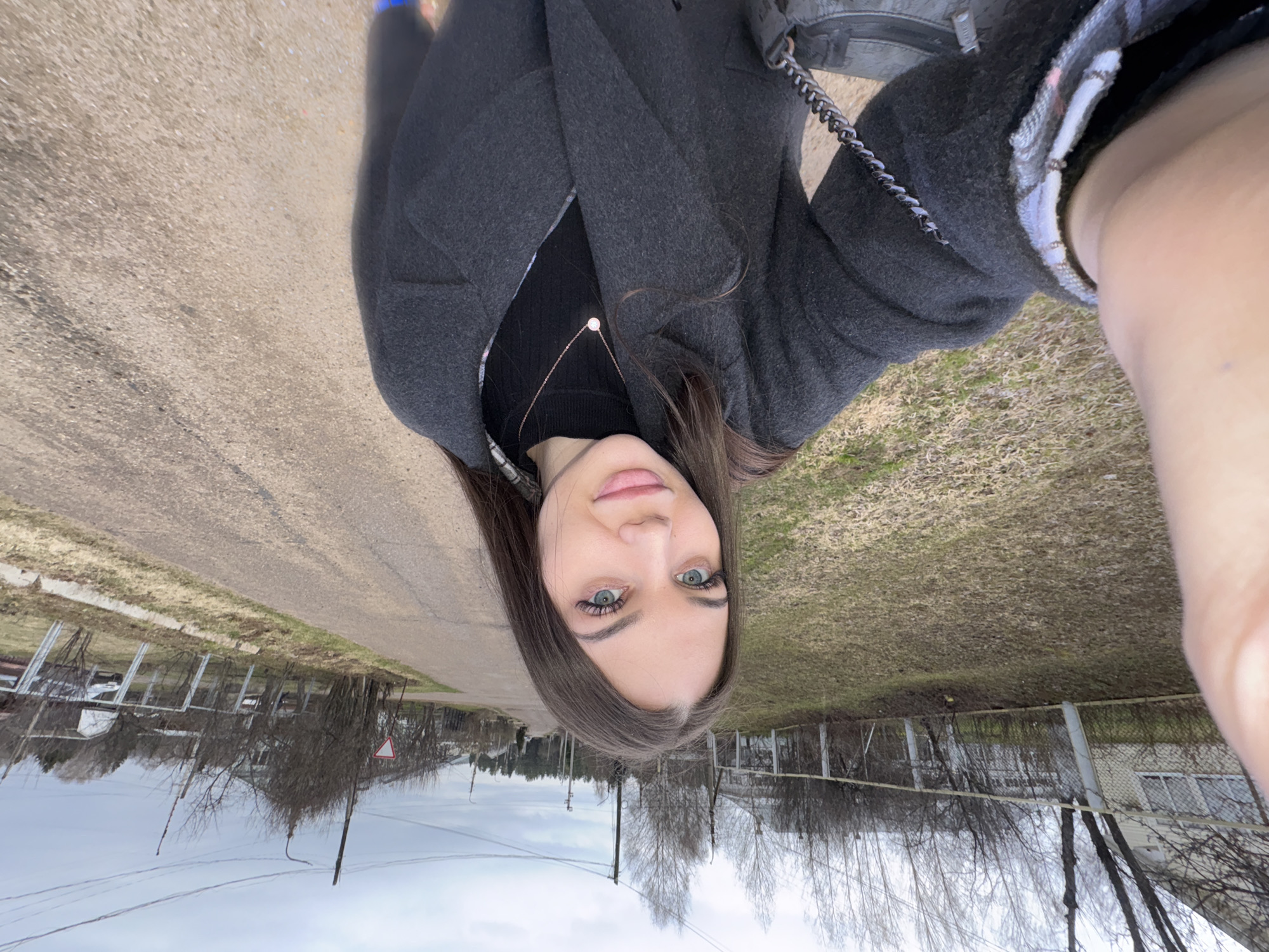 A selfie taken in Прикамский, Russia, shows a young woman with long brown hair, light skin, and light blue eyes. She wears a dark gray coat, and a black top with a simple silver necklace. The photo is taken in a residential area, showing a street with a bare tree lined median, a sidewalk, bare trees and overcast skies. There is a building with a wire fence nearby.