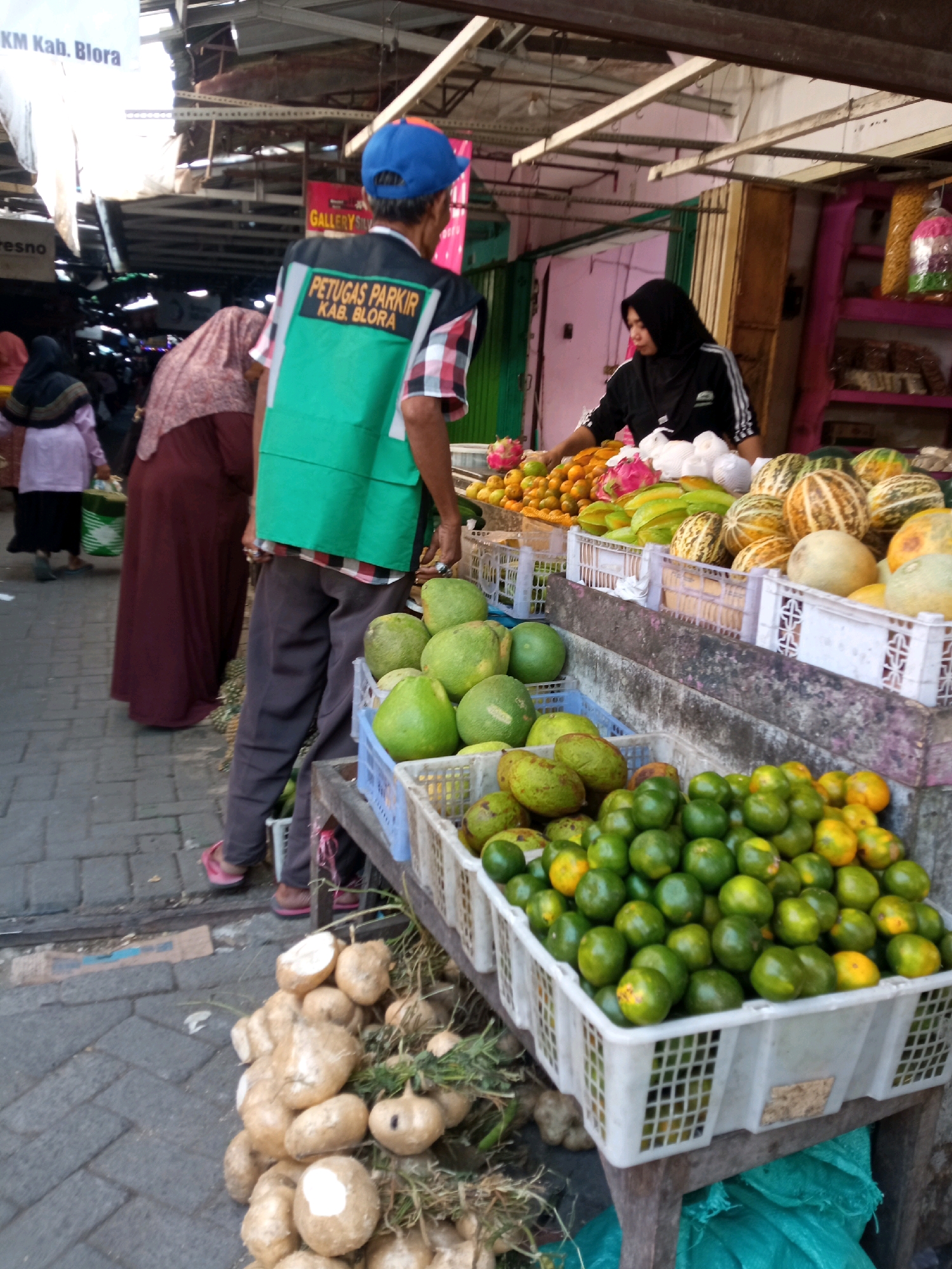 The image depicts a bustling market scene, likely in Balun, Indonesia. In the foreground, a person wearing a green vest with "PETUGAS PARKIR KAB. BLORA" (Parking Officer Kab. Blora) printed on it stands next to a display of produce. The vest is a bright green with orange text. The individual is wearing dark pants and pink sandals. Behind this person, other market-goers are visible, including a woman in a dark hijab and a maroon-colored abaya.
The market stall features an abundance of fresh fruits and vegetables. Baskets are filled with what appear to be oranges, limes, melons, and possibly dragon fruit. Some of the melons have a distinctive striped pattern. In the lower left of the image, a pile of root vegetables, possibly jicama, is visible. The ground is paved with interlocking gray bricks. In the background, stalls and awnings suggest a covered market area, with some goods displayed on shelves. A sign with "GALLERY" is partially visible. The overall impression is one of a vibrant and active local marketplace.