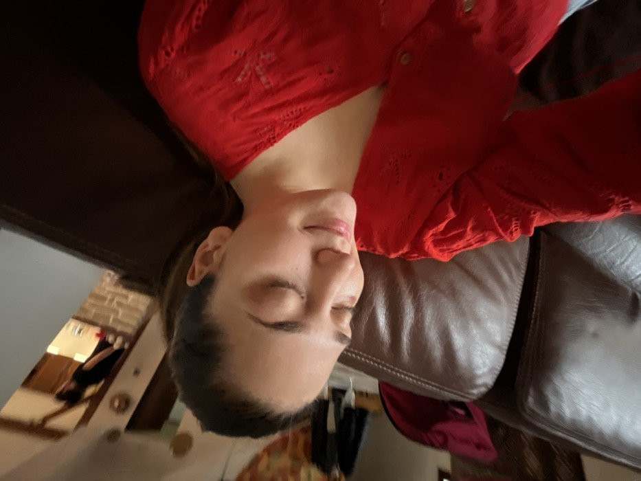 The scene is located in a residence in Ancenis, France. A young individual with dark hair, pulled back from the face, is depicted lying on their left side on a brown leather sofa. Their eyes are closed, and a slight smile is visible. They are wearing a long-sleeved red top with a textured or patterned fabric. In the blurred background, another person, dressed in a dark top, is partially visible within an adjacent, brightly lit indoor area. This background space suggests a domestic setting, possibly a kitchen or dining area, with light-colored walls and a tiled surface visible.