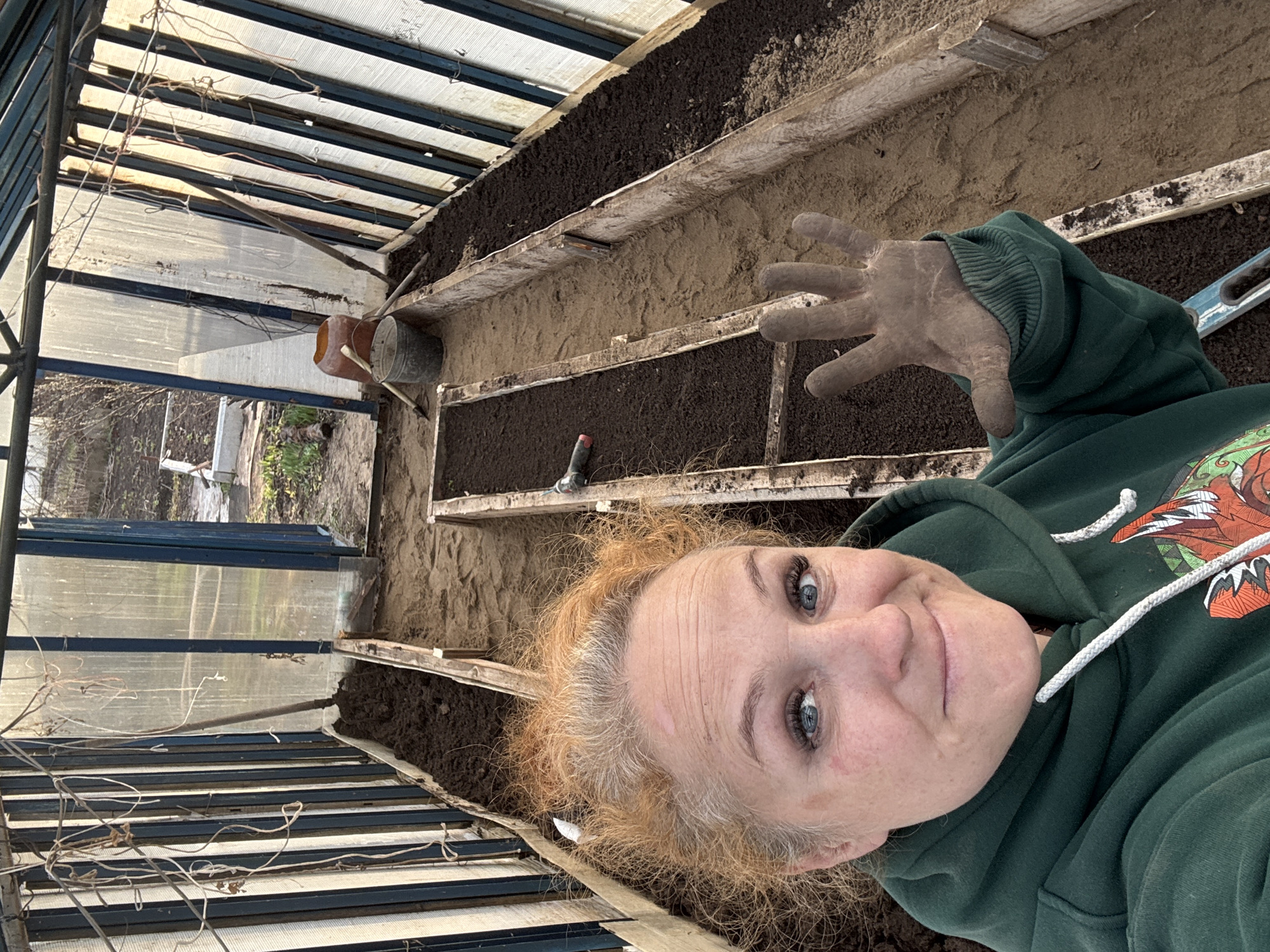 A selfie shows a woman inside a greenhouse. The woman, wearing a green hoodie, is in the foreground, smiling with her blue eyes visible. Her hair is fair. She is wearing a gardening glove on one hand. In the background, the greenhouse's interior features rows of raised garden beds filled with dark soil. A watering can and small gardening tools are visible. The greenhouse's structure has transparent panels and is located outdoors, with some foliage visible outside. The location is Chaikovsky, Russia.