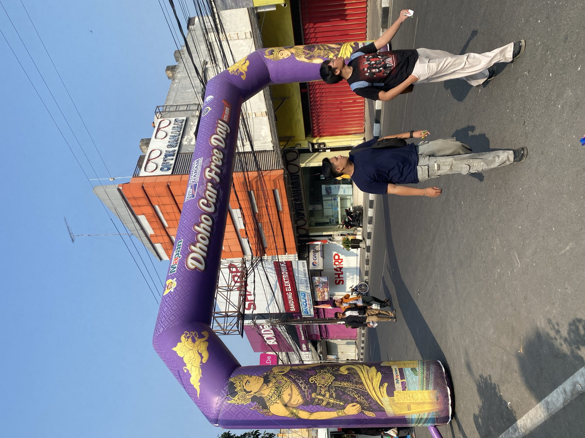 This image captures a "Dhoho Car Free Day" event in the city of Kediri, Indonesia. A large, inflatable purple archway stands prominently in the foreground, decorated with traditional Javanese motifs and figures. The text on the arch reads "Dhoho Car Free Day" and includes branding for "Mapans" and "Gudang Garam".
In the middle ground, two young men are walking towards the viewer. The man on the left wears a dark blue t-shirt and grey cargo pants, carrying a black bag. The man on the right wears a black t-shirt with a graphic print and light-colored pants, also carrying a bag. Several other individuals are visible walking in the background, some near a white tent and a bicycle.
The backdrop consists of buildings lining the street. One building features an orange facade with signage that includes "OPTİK SAHABAT". Another building displays "BOLDEX" and "BANDUNG ELEKTRONIKA". Various overhead wires and antennas are visible against a clear blue sky, indicating an urban environment. The street itself is asphalted, marked with white lines, and shows dappled sunlight and shadows. The overall scene depicts an active community event in a commercial area.
