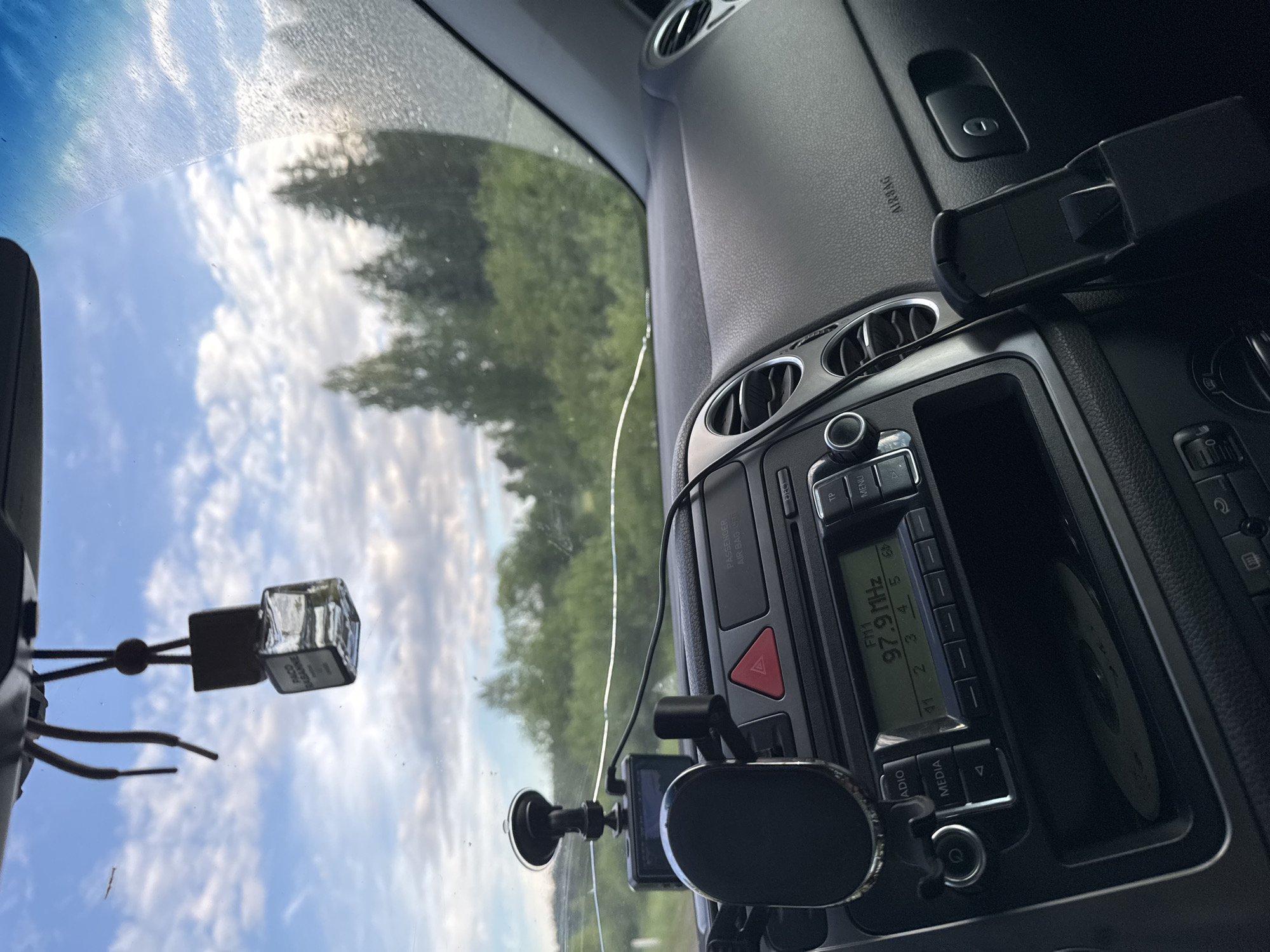 The image shows the interior of a vehicle. The dashboard and windshield are prominent. A radio display shows a frequency of 97.9 MHz. There are dashboard controls, air vents, and an airbag indicator. An air freshener hangs from the rearview mirror. A smartphone holder is attached to the windshield. Through the windshield, trees and the sky are visible. It is not possible to determine the location as Malakhovo, Russia from the image.
