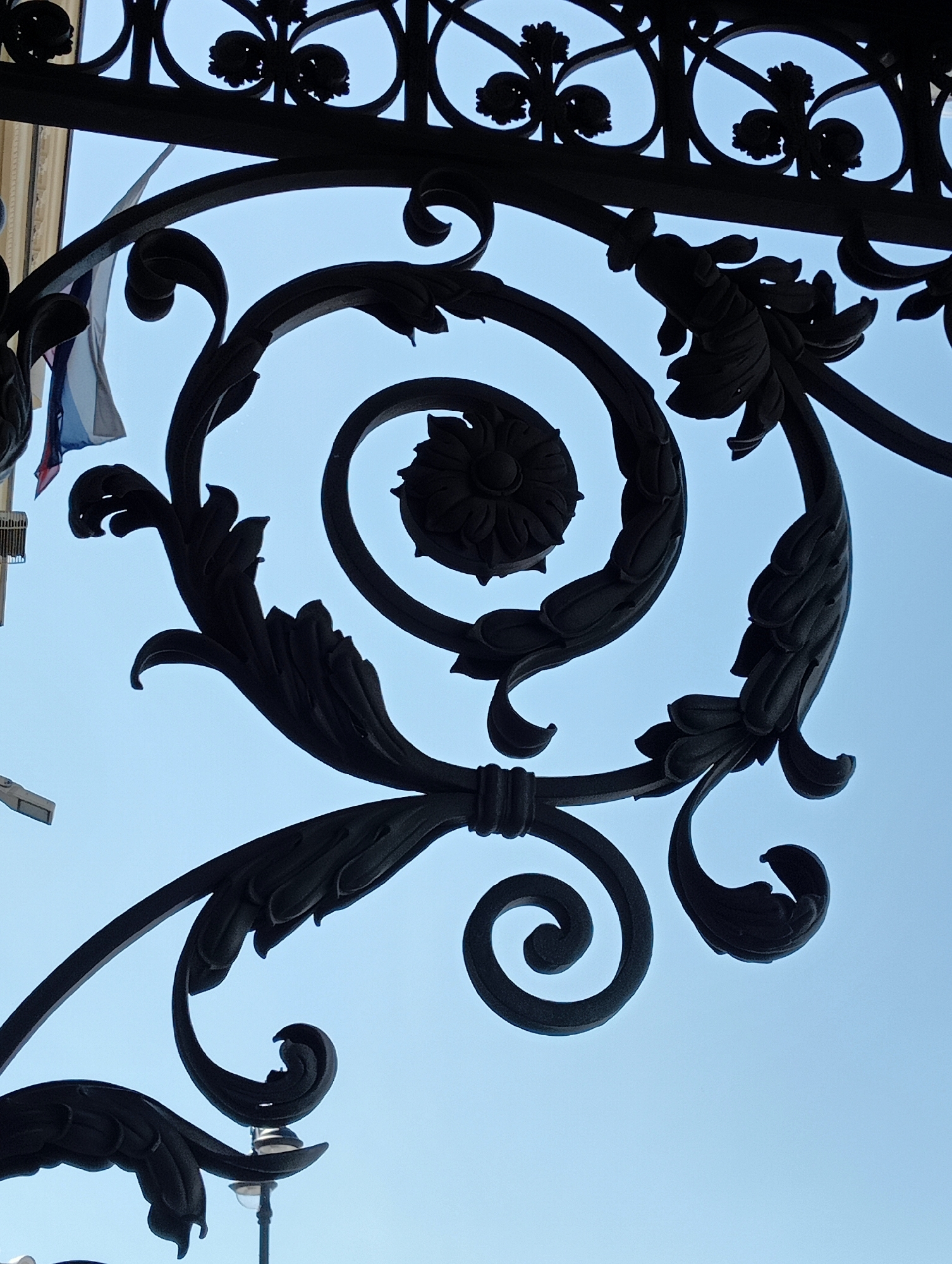 The image shows a close-up, low-angle shot of ornate black wrought ironwork against a clear blue sky. The ironwork features intricate scrolling, leaf-like motifs, and floral designs. In the upper left, a small section of a flag with vertical stripes of white, blue, and red is visible, suggesting the location is likely Russia. While not explicitly identifiable, the architectural style of the surrounding buildings, implied by the glimpses in the cropped hints, and the presence of the Russian flag strongly point to a location in Saint Petersburg, Russia. The scene is static, with no people or actions depicted, focusing solely on the detailed craftsmanship of the ironwork and the contrast with the sky.