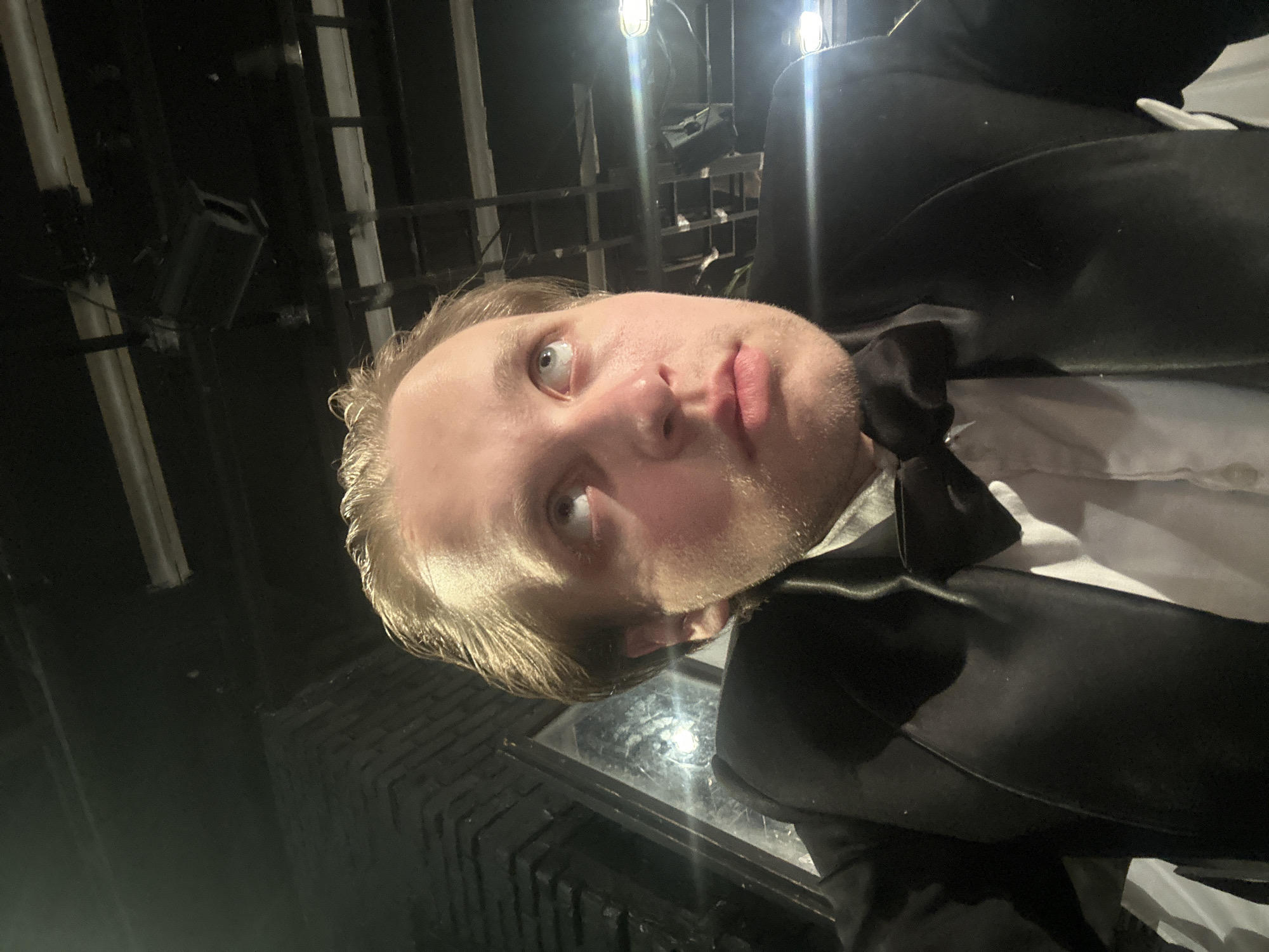 The image shows a close-up, low-angle selfie of a young Caucasian male with blond hair and light-colored eyes. He is wearing a black tuxedo jacket, a white dress shirt, and a black bow tie. His head is tilted upwards, and his gaze is directed towards the ceiling. The background appears to be an indoor, dimly lit environment, possibly a theater or backstage area, with exposed ceiling structures and lighting fixtures visible. Some reflections are present on a glass-like surface to the left of the subject. The overall lighting is dramatic, with strong highlights on the subject's face. The location is identified as Unknown, Unknown.