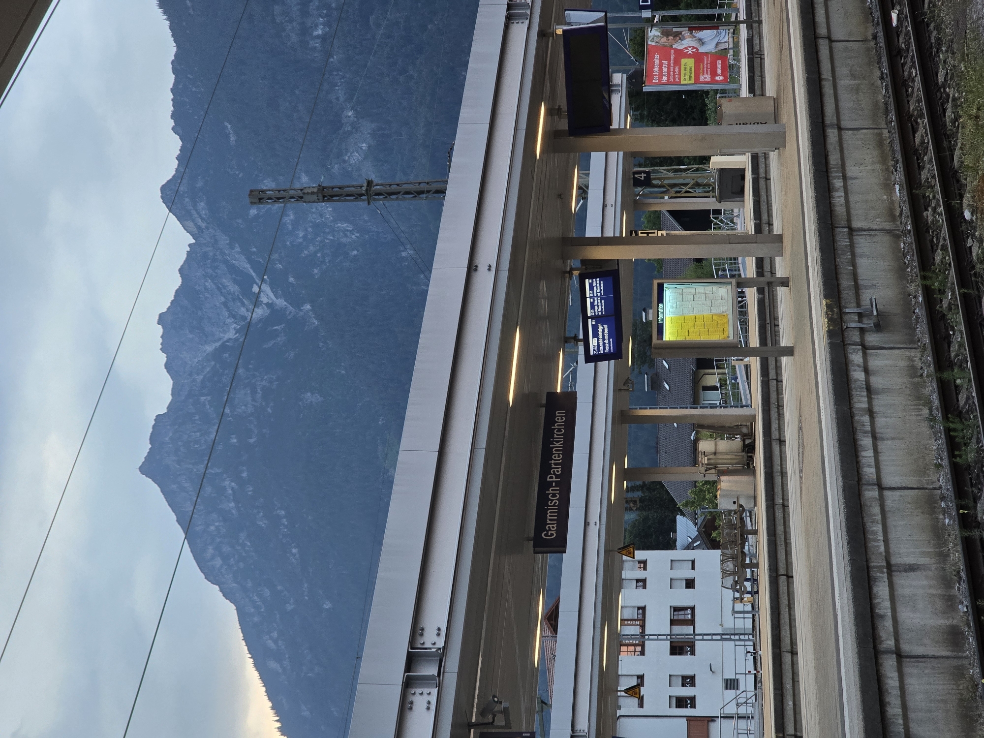 The image captures a scene at the Garmisch-Partenkirchen train station in Germany. In the foreground, train tracks are visible, leading towards a concrete platform. The platform itself is covered by a modern, multi-level roof structure supported by metal pillars. A prominent sign displaying "Garmisch-Partenkirchen" in dark letters on a lighter background hangs from the roof.
To the left of the platform, a white building with multiple windows is visible. Scattered across the platform are several empty benches and some trash receptacles. To the right, there are electronic display boards, one of which shows train departure information with the text "Bitte nicht einsteigen Please do not board" and departure times. Further to the right, a large red advertisement poster is mounted on a pole.
In the background, a rugged, tree-covered mountain range dominates the skyline under a cloudy sky. The overall impression is that of a functional, well-maintained train station situated in a scenic mountain environment. No people are clearly identifiable in the image.