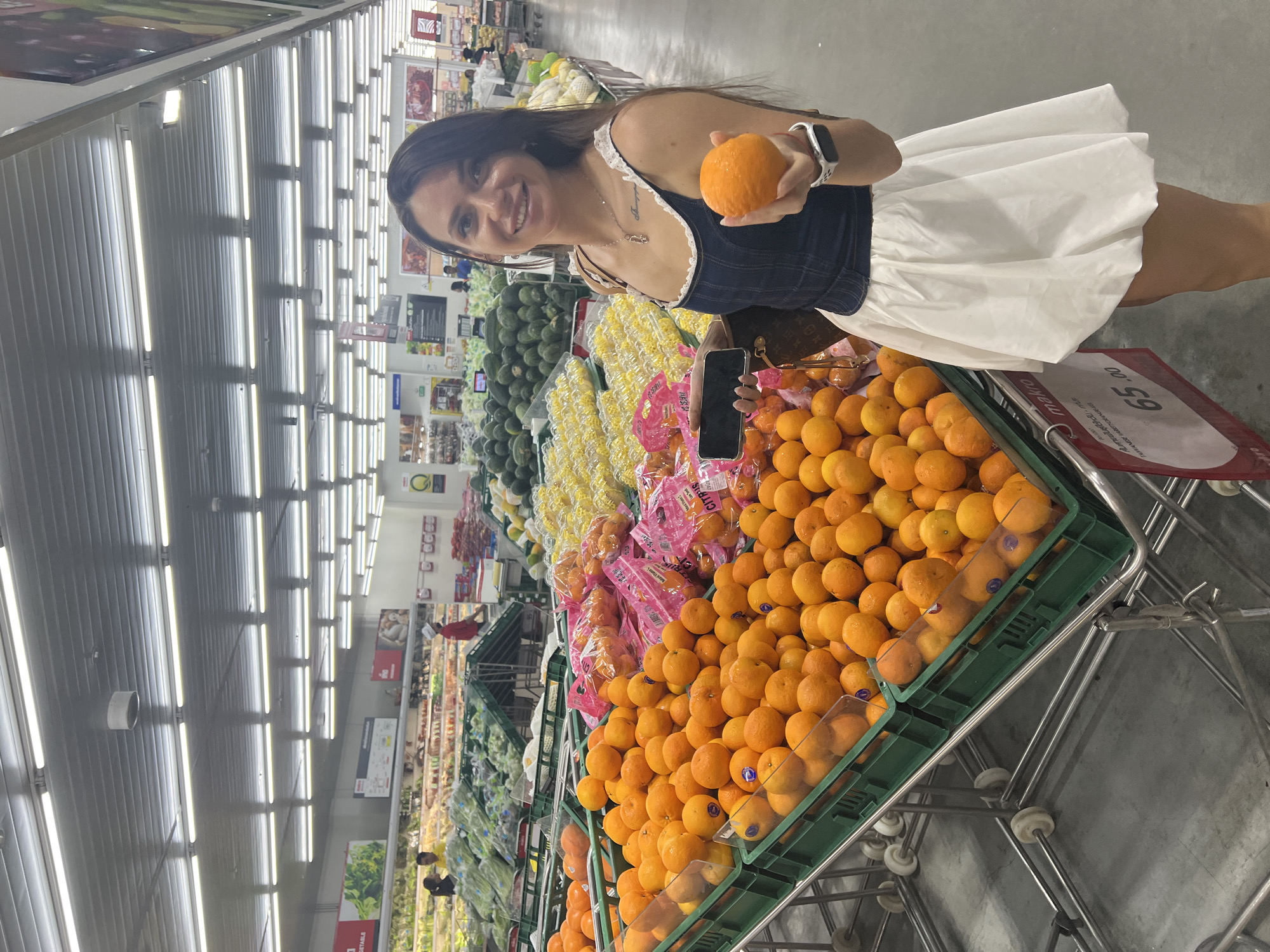 In a brightly lit aisle of a grocery store in Baan Hua Thanon, Thailand, a woman is standing near a display of oranges. She is holding an orange in her right hand and a mobile phone in her left. The woman is smiling. The grocery store has rows of produce including avocados, corn, oranges, and other fruits and vegetables. Fluorescent lights illuminate the aisle from above. The price of the oranges is displayed on the cart, showing 65.00.