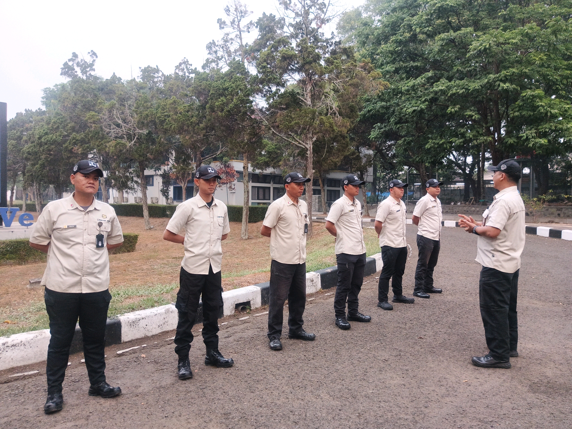 Six men in uniform stand in a line on a paved surface, with trees and a building in the background. The men are wearing tan collared shirts, black baseball caps with logos, and black pants. Five of the men are facing forward, while one man on the right is facing left and gesturing with his hands. The man on the far left is closest to the camera, and the men are spaced out evenly. The building in the background appears to be a low-rise structure with windows. The environment suggests an outdoor, possibly institutional or commercial setting.